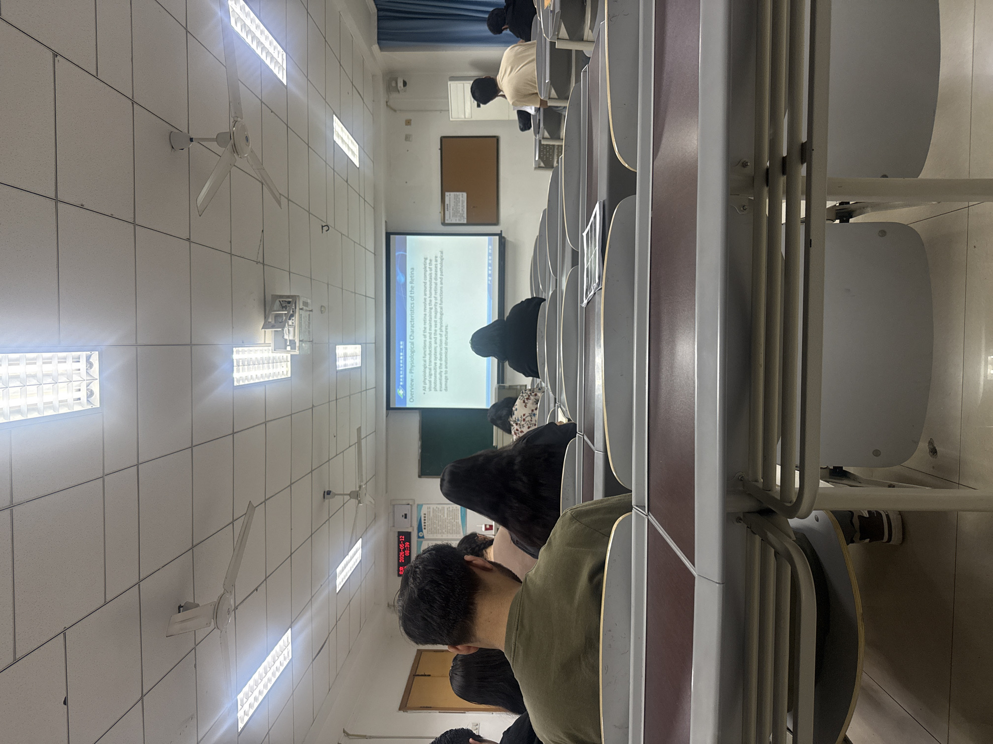 The image shows a classroom setting, likely in Yuzhong District, China, based on the on-screen timestamp. Rows of students sit at desks facing a projector screen displaying a lecture slide about the "Physiological Characteristics of the Retina." A few students are visible, including one in a green shirt in the foreground, and others further away with black clothing. The classroom has fluorescent lights, ceiling fans, a bulletin board, and air conditioning units on the walls. The focus is on the lecture and the student's perspective from within the classroom.