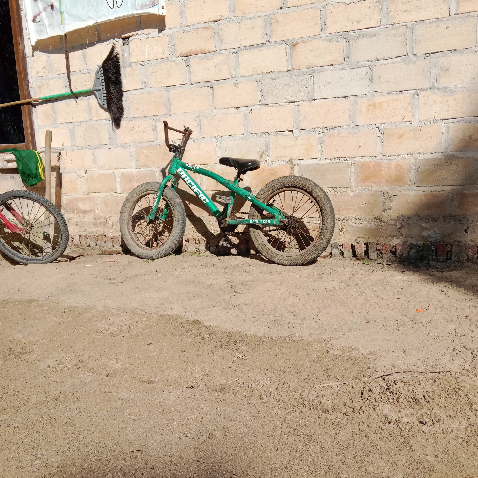 In a dusty, outdoor setting against a light-colored brick wall, a green BMX bicycle is parked. The bicycle, which appears to be for children or a smaller rider, is leaning against the wall. It has a black seat, handlebars, and thick tires. The word "PACIFIC" is visible on the frame, along with "COOL TECH" on the lower frame. To the left of the BMX, the front wheel of another bicycle with a red frame is visible. A broom with a green handle and a black brush is propped against the wall to the left of the bicycles, near a window with a wooden frame. A piece of green fabric with yellow trim is hanging on the wall to the left of the broom. The ground is made of packed dirt with some scattered debris. The scene suggests a utilitarian outdoor area, possibly a yard or alley, in a residential setting.