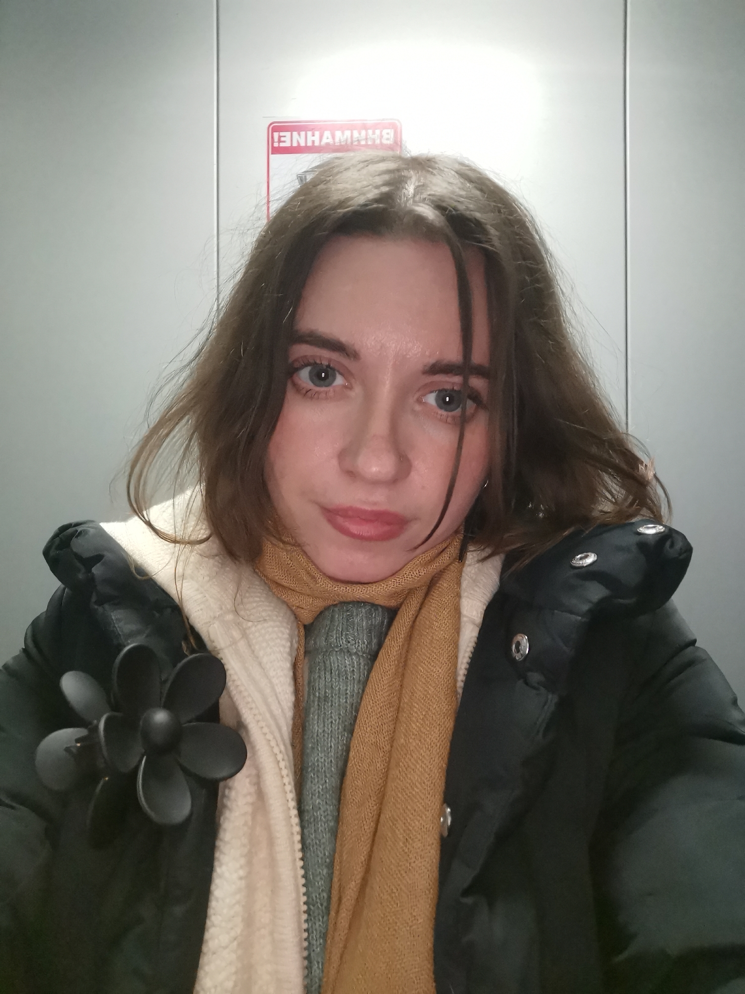 A young woman is taking a selfie in what appears to be an elevator. She has light skin, blue eyes, and light brown hair. She is wearing a black puffer jacket over a cream-colored hooded sweater, a gray sweater, and a mustard-yellow scarf. A large, black flower pin is attached to her jacket. The elevator has gray walls and a red and white sign is visible in the background. The location is unknown.