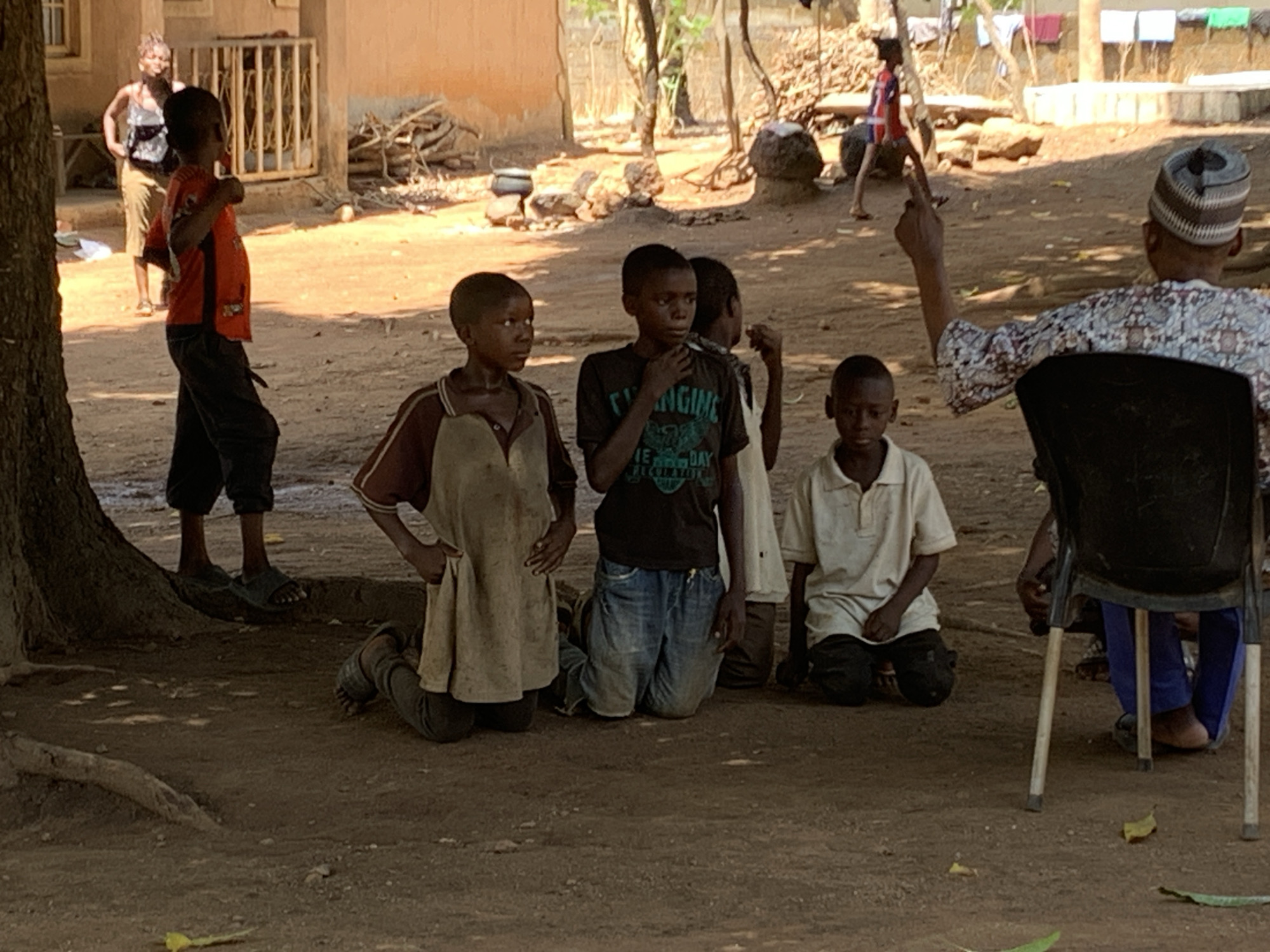The image shows a group of four young boys kneeling on the ground in what appears to be a village setting in an area identified as Unknown, Unknown. One boy has his hand on his chin. A man, partially visible from the back, sits on a plastic chair nearby, with his right arm raised and index finger pointing upwards. Another boy stands to the left, wearing an orange shirt. In the background, there's a woman in a tank top, standing next to a building, and another person further away. The setting has a dirt ground with trees and scattered items, suggesting a rural environment. Clothes hang on a line in the background, and there's what appears to be a wood pile near the building.