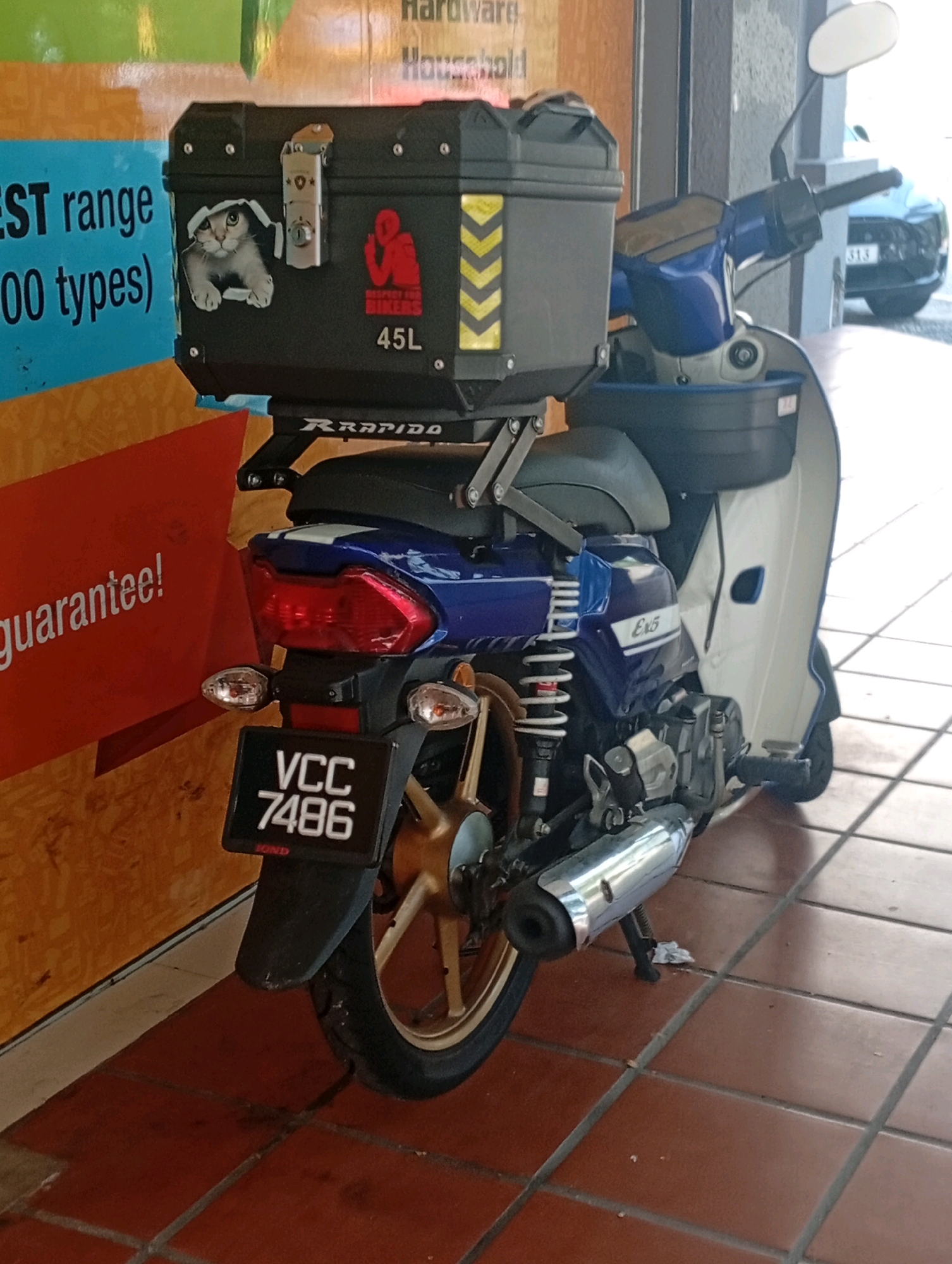 A blue and white scooter is parked on a tiled floor, viewed from the rear three-quarter angle. The scooter is equipped with a large black storage box on the rear rack, which has a sticker of a cat peeking out from under a blanket, a "Respect for Bikers" logo, and "45L" printed on it. A silver lock is attached to the top of the storage box. The scooter's license plate reads "VCC 7486". To the left of the scooter, a portion of a poster with text about "EST range" and "00 types" is visible. To the right, a black car with license plate "313" is parked in the background. The setting appears to be an outdoor area with a tiled floor and a wall with colorful posters.