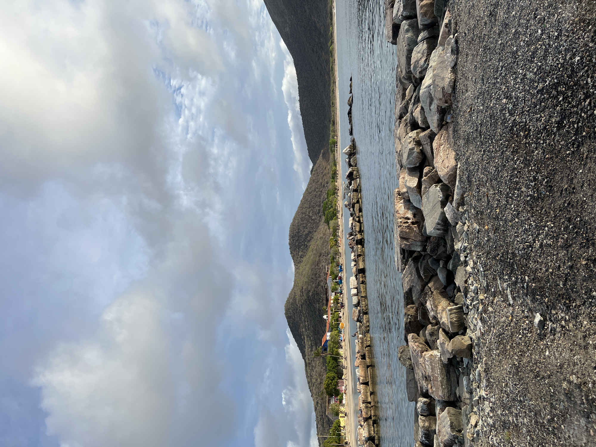 The image shows a waterfront scene in Pedro González, Venezuela. In the foreground, a rocky shore and a breakwater made of large stones line the edge of the water. The water itself is calm, with gentle ripples reflecting the sky. Beyond the breakwater, a sandy beach is visible, with a line of buildings situated behind it. These buildings appear to be a mix of houses and possibly small businesses, with some featuring colorful facades. Green trees and vegetation are scattered along the beach and around the buildings. In the background, two prominent, rounded hills rise above the landscape, covered in dry, scrubby vegetation. The sky above is partly cloudy, with dramatic cloud formations casting shadows and allowing sunlight to break through in places. The overall scene depicts a tranquil coastal environment with evidence of human settlement.