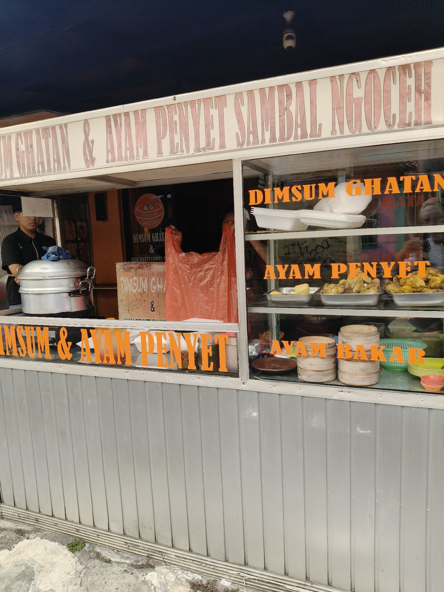 The image shows a food stall in South Tangerang, Indonesia, specializing in "Dimsum Ghatan & Ayam Penyet Sambal Ngoceh." The stall has a prominent sign above the serving area. Inside, a person wearing a black shirt is visible behind the counter, near a large silver steaming pot. Orange plastic bags and various food items, including what appear to be dim sum and fried chicken, are displayed on shelves within a glass-enclosed serving window. There are stacks of bamboo steamers and bowls on the counter. The exterior of the stall features a metallic, corrugated metal paneling at the bottom. The overall impression is of a busy street food vendor.