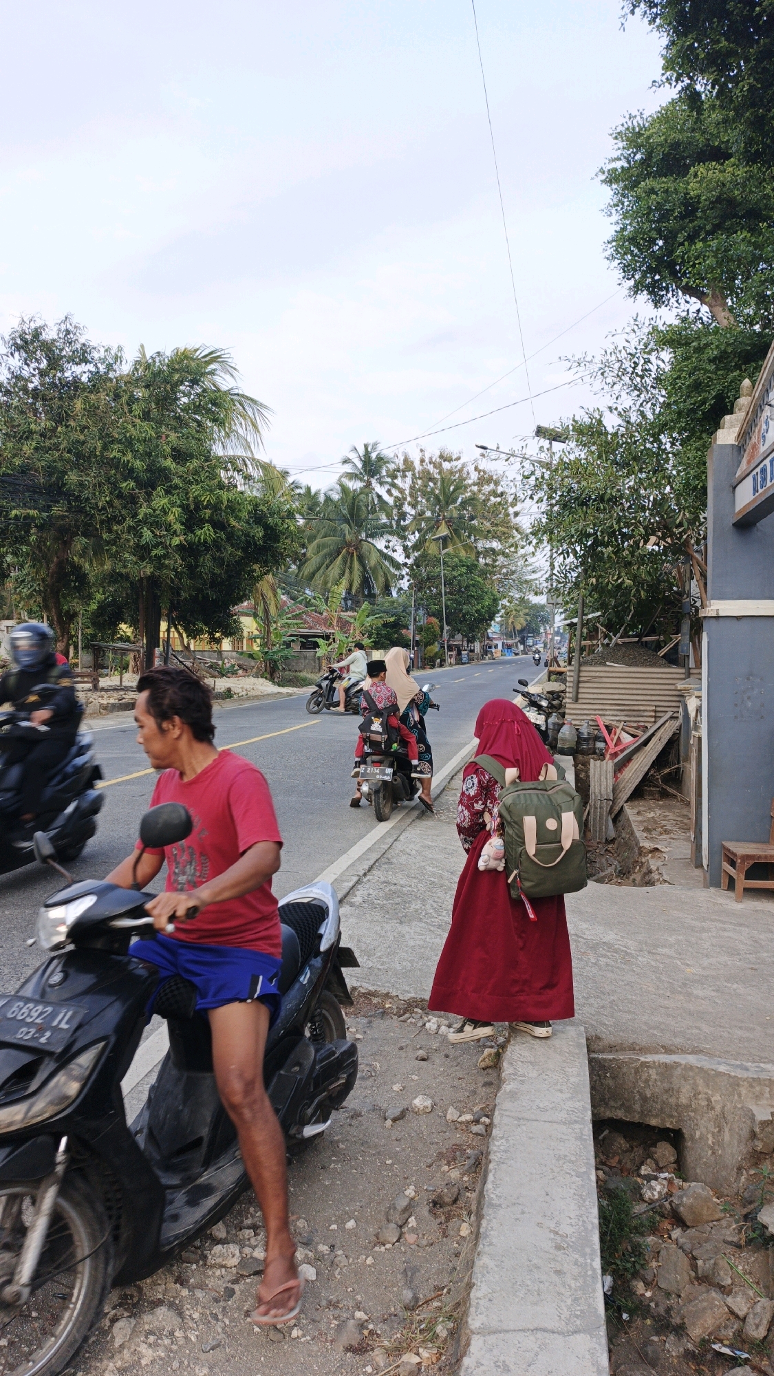 A candid, full-shot, eye-level, outdoor, daytime scene captures a road in Cibenda, Indonesia. A man in a red t-shirt and blue shorts sits on a black motorcycle. A woman in a red hijab and a floral-patterned top stands on a concrete sidewalk, carrying a green backpack. Another woman on a motorcycle, wearing a beige hijab, is in the background. The road is lined with trees and palm trees. The sky is overcast.