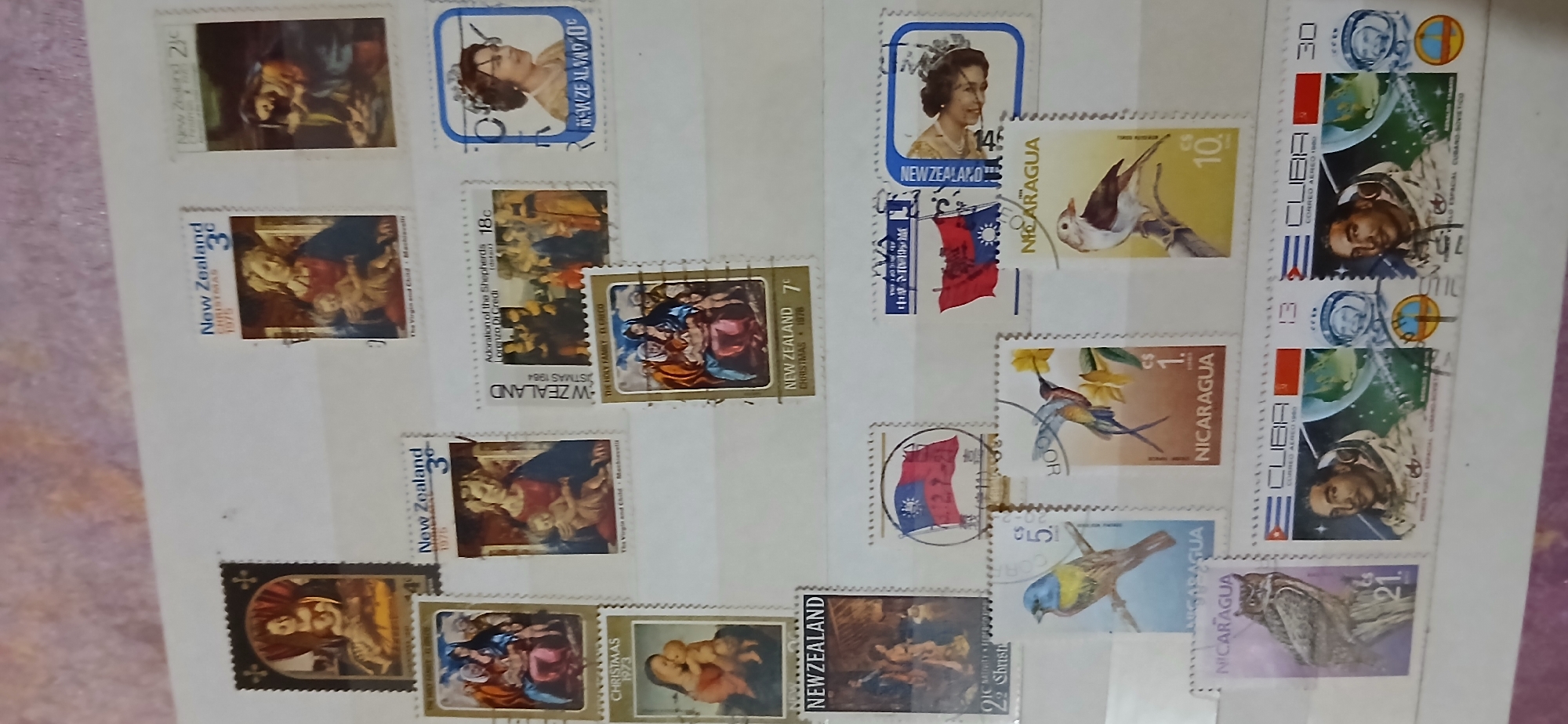 The image shows a collection of postage stamps arranged on a white surface, likely an album page. The stamps are of various countries, including Nicaragua, Cuba, New Zealand, and Taiwan. The designs feature a range of subjects such as birds, astronauts, religious scenes (depicting the Virgin Mary and child, and the Annunciation), and national flags. The stamps are in different sizes and colors, with perforations along their edges. The lighting suggests a bright, possibly natural light source. There is no indication of the city of Samarinda, Indonesia in the image.