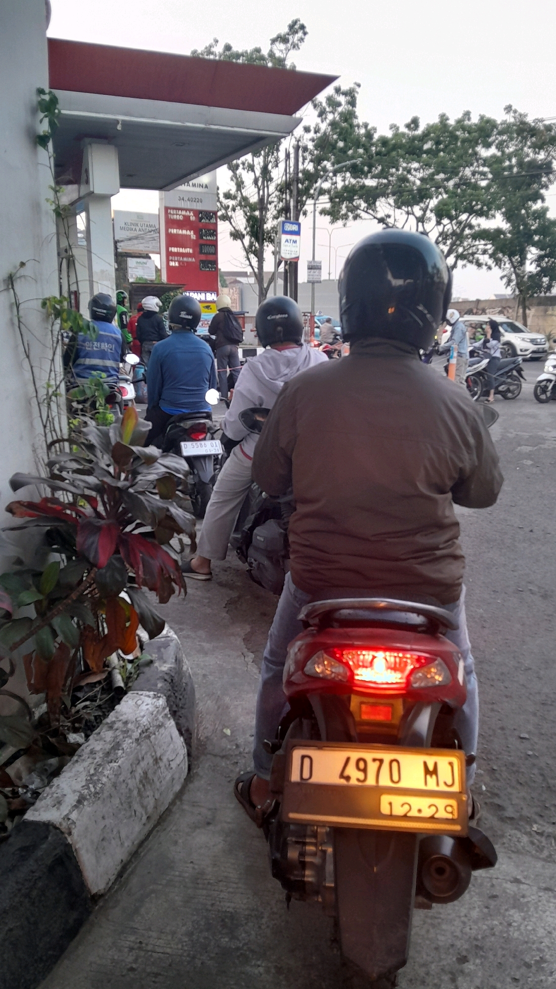 In an urban outdoor scene, several people on motorcycles are lined up at a gas station, with a red canopy overhead. The time of day appears to be late afternoon or early evening, given the lighting conditions. On the left side of the image, a plant with dark red and green leaves grows near a concrete curb. The gas station has signage indicating fuel prices and an ATM. The motorcycles are diverse in color, including a red one in the foreground with a license plate that reads "D 4970 MJ" and "12.29". Most of the people are wearing helmets and casual clothing. The environment includes a paved road and trees in the background.