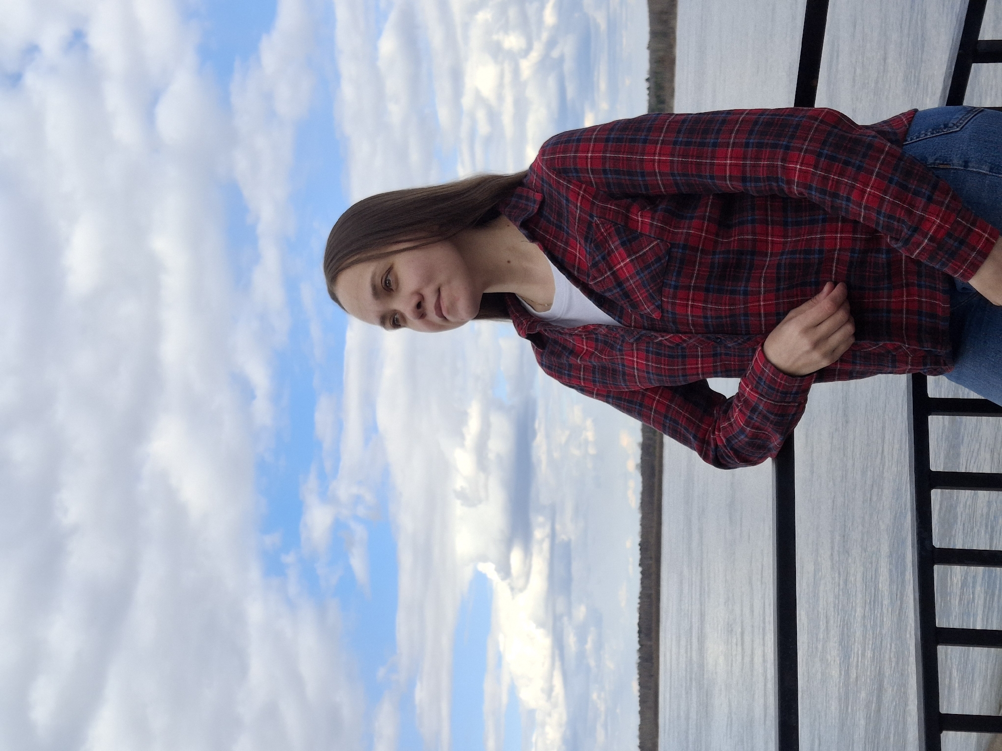 A young woman with long, straight brown hair is depicted standing outdoors, looking slightly off-camera to her left with a neutral, possibly thoughtful, expression. She is dressed in a red and black plaid flannel shirt worn open over a white t-shirt, and blue jeans. Her left hand is resting near her waist.
The background features a large body of water, likely a river or lake, extending to a distant, dark horizon line that suggests a far shore or forest. Above the water, a bright, partly cloudy sky fills the upper part of the frame, showing a mix of substantial white and grey clouds and patches of clear blue. A black metal railing or fence is visible in the foreground on the right side, indicating the woman is on a platform, pier, or bridge overlooking the water.
The setting appears to be daytime, with soft, diffused lighting under the cloud cover. The scene evokes a calm and tranquil atmosphere. Based on the provided context, the image is set in Sarapul, Russia, suggesting the prominent body of water is the Kama River. No visible text is present in the media.