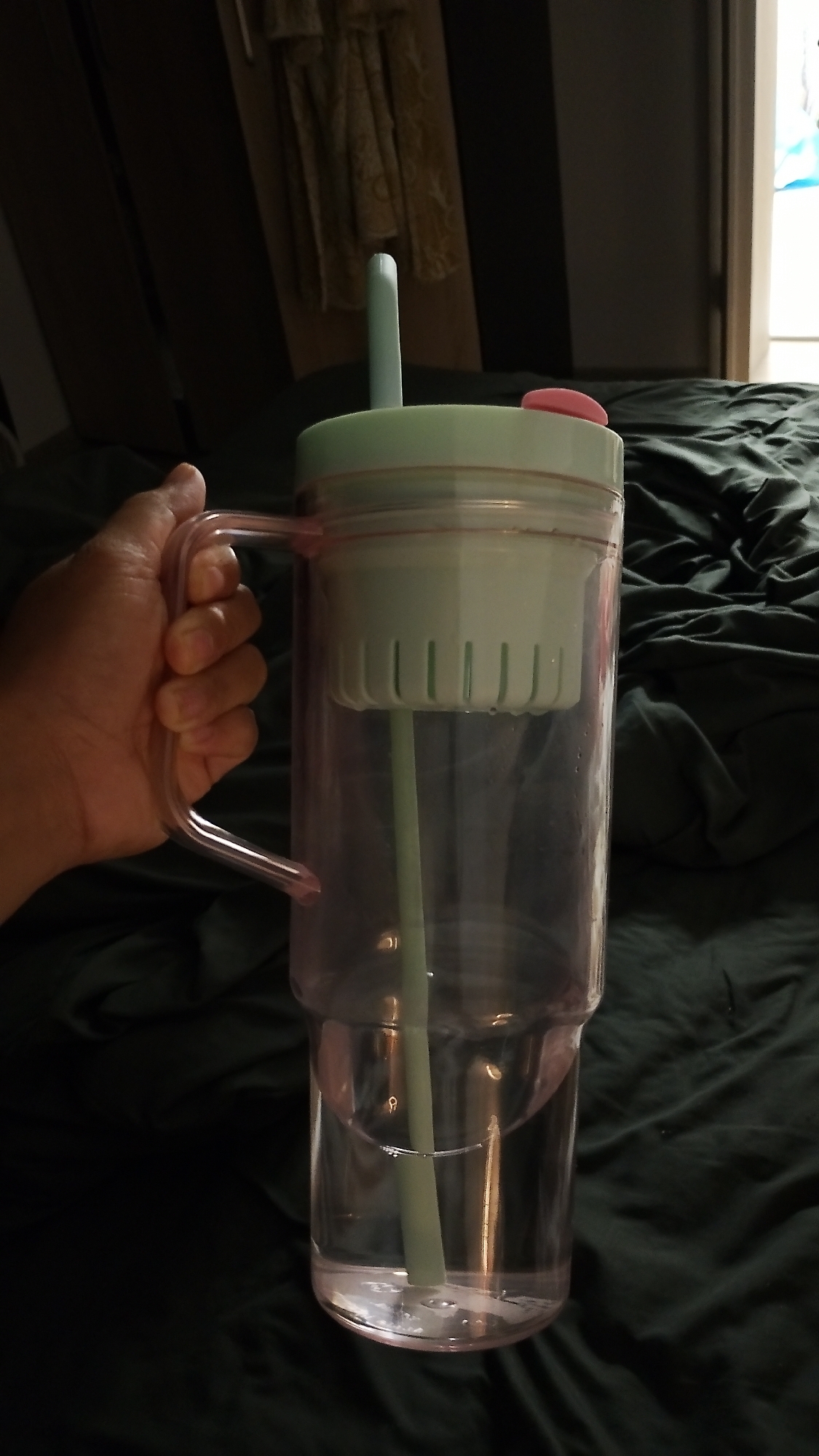 A hand holds a large, translucent pink plastic tumbler with a light green lid and straw. The tumbler is about two-thirds full of clear liquid. The lid has a small pink button on top. The background consists of dark green, rumpled fabric, likely bedding, and a partially visible window with blinds and curtains. The image is taken indoors and is dimly lit. No information about Smolyan, Bulgaria is discernible from the image.