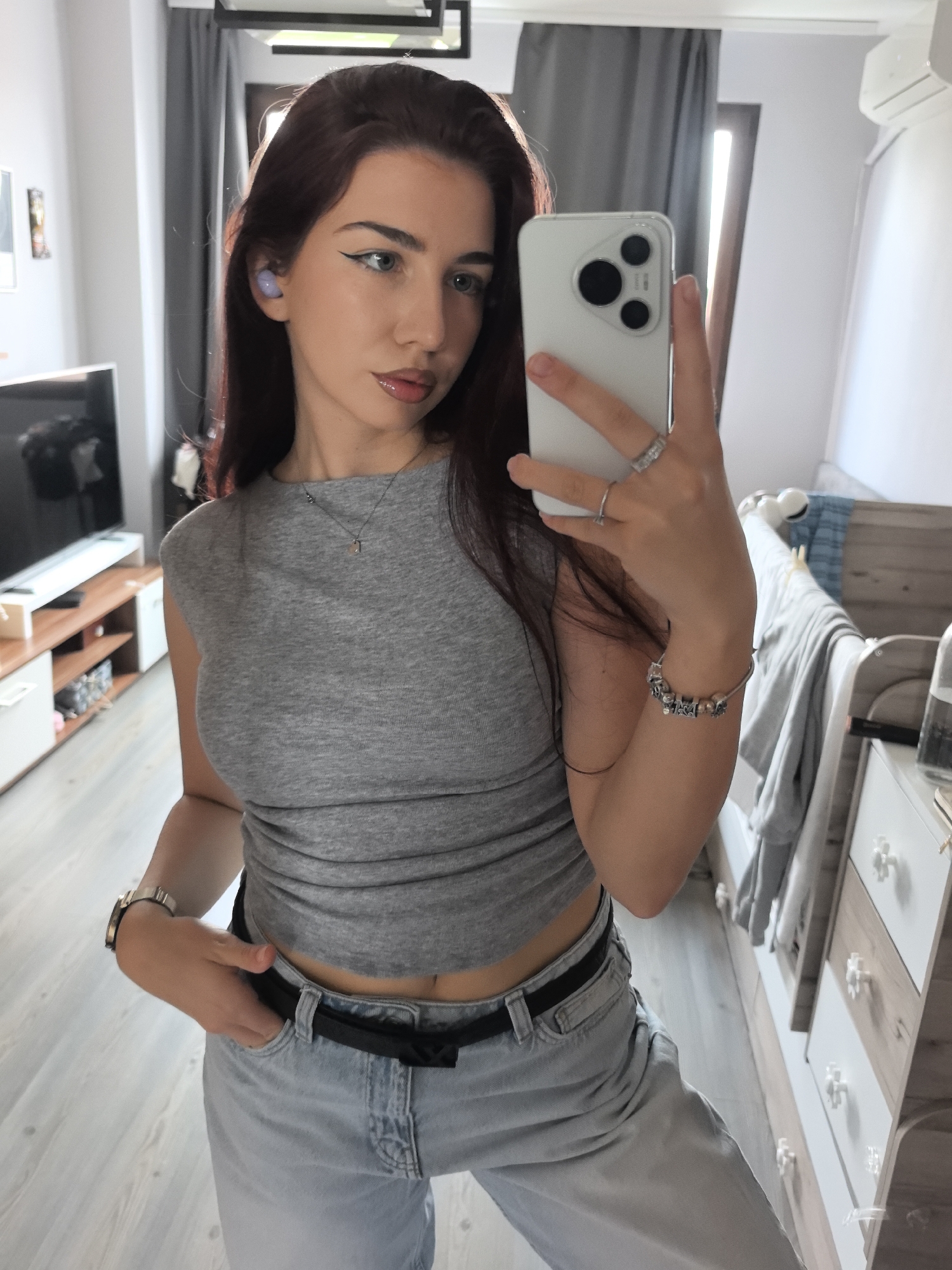 A young woman is captured in a mirror selfie inside what appears to be a modern home or apartment in Stara Zagora, Bulgaria. She has long, dark reddish-brown hair and is wearing a grey sleeveless crop top, light blue/grey jeans, and a black belt. Her left hand is casually placed in her jeans pocket, while her right hand holds a white smartphone with a triple-lens camera setup, used to take the photograph.
Notable accessories include a silver watch on her left wrist, a silver necklace with a small pendant, multiple silver rings on her right hand, and a silver charm bracelet on her right wrist. A light purple wireless earbud is visible in her left ear. She has prominent winged eyeliner and glossy lips, maintaining a neutral to slightly contemplative expression as she looks into the mirror.
The indoor setting features a light wooden floor and white walls. In the background, to the left, there's a wooden TV stand with white drawers, holding a large, off flat-screen television. To the right, a white and wooden dresser with decorative clear handles is visible, with a bed frame partially seen behind it. Natural daylight streams into the room from a window or door covered by dark grey curtains in the background, suggesting it is daytime. A black rectangular light fixture is mounted on the ceiling. No discernible text is visible within the image.