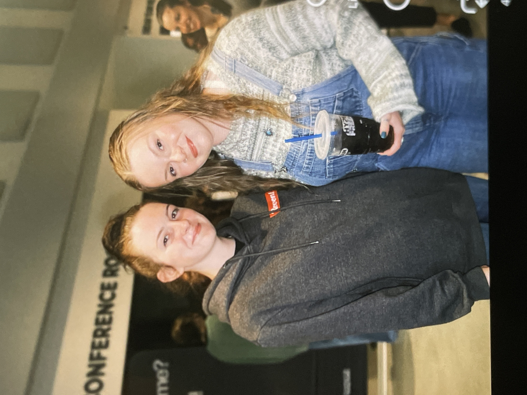 Two young women pose for a picture in an indoor setting. The woman on the left has her hair in a bun and is wearing a dark grey hoodie. The woman on the right has long, wavy blonde hair and is wearing denim overalls over a patterned sweater. She is holding a clear plastic cup with a dark beverage and a blue straw. In the background, a sign reads "CONFERENCE RO." The overall lighting is dim, suggesting an indoor event or venue.