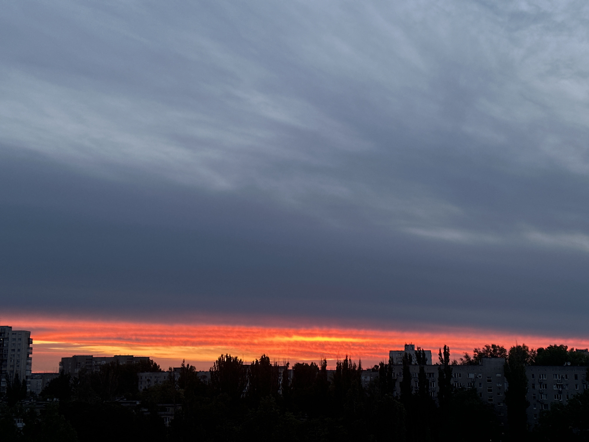 The image captures a cityscape at dusk or dawn in an urban area of unknown location. The sky is predominantly a gradient of dark grey to light grey with horizontal streaks of clouds. A vibrant band of red and orange light illuminates the horizon where the sky meets the city skyline.
The cityscape features a row of buildings with varying heights and architectural styles. Silhouettes of trees are visible in the foreground, adding depth to the scene. The image appears to be taken from an elevated position, possibly a rooftop or upper-story window, providing a panoramic view of the city.