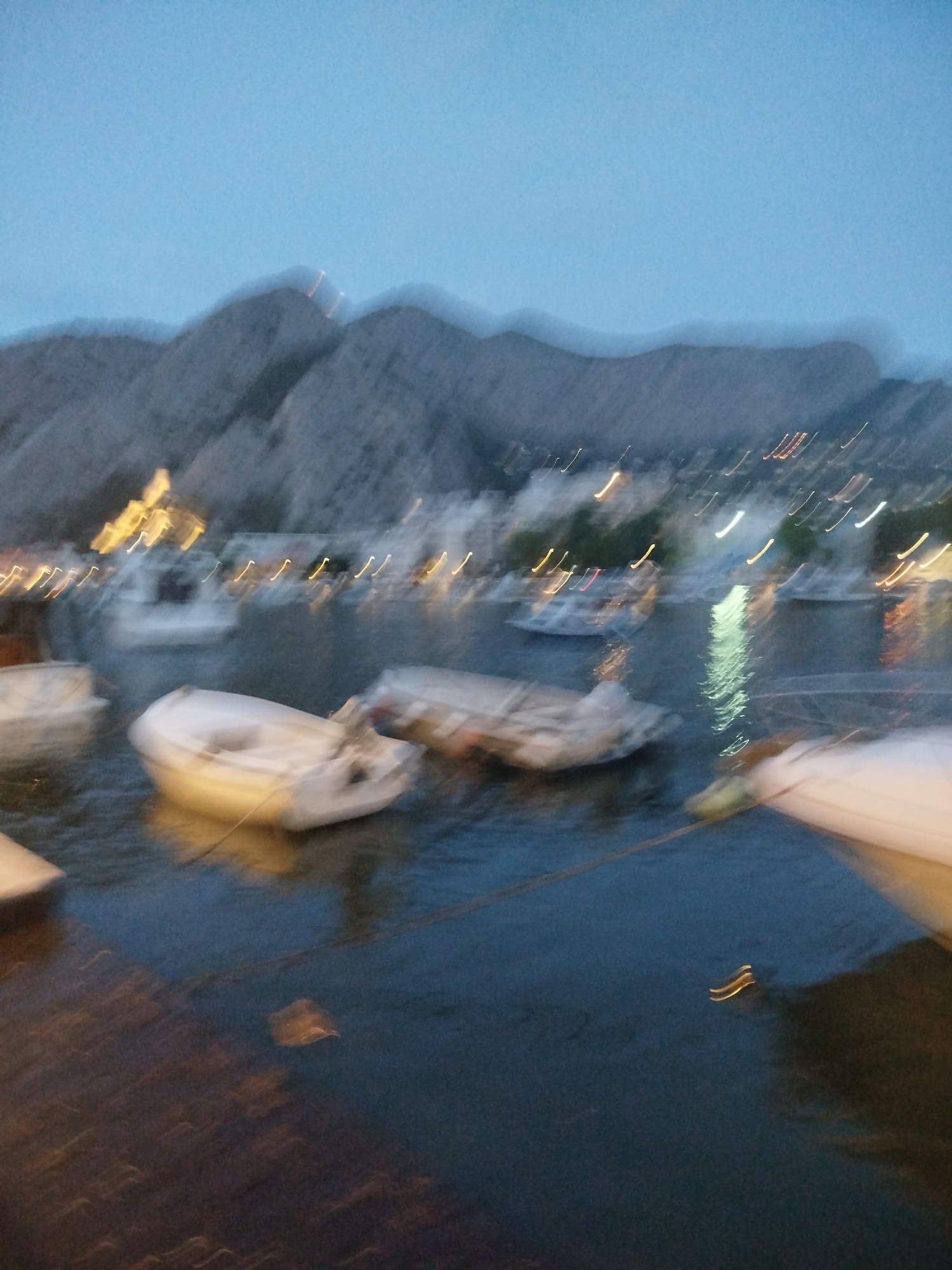 The image depicts a harbor at dusk or dawn, with a town nestled at the base of a mountain range. The scene is characterized by motion blur, suggesting the photograph was taken with a slow shutter speed or from a moving platform. Several small boats are visible in the water, some appearing to be dinghies or small fishing vessels. The lights of the town are reflected in the water, creating streaks of light. The sky is a pale blue, transitioning into darker hues as it nears the horizon. The mountain range in the background is indistinct due to the blur, but appears to be rugged and mountainous. The foreground shows a portion of a wooden dock or pier. The overall impression is one of a tranquil, waterside location at the edge of a mountainous region, likely a coastal town. Given the visual cues of the steep mountain backdrop and the type of harbor, this scene is likely located in Omiš, Croatia, a town renowned for its dramatic mountainous setting and its position at the mouth of the Cetina River.