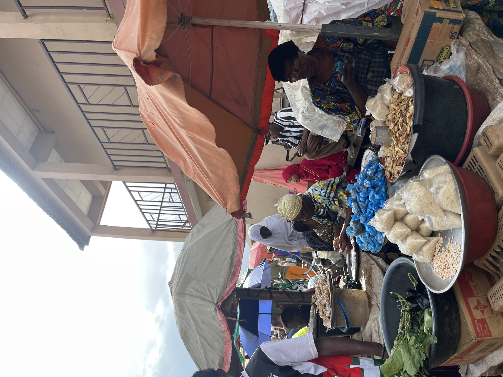 This media file captures a vibrant scene at an outdoor market in Rafin Par, Nigeria. Several people are visible, seemingly engaged in commercial activities. The subjects include at least three women and a few other individuals whose faces are partially obscured. One woman, wearing glasses and a colorful patterned top, appears to be interacting with a customer or vendor. Another woman is dressed in a red hijab and a matching garment, while a third wears a white head covering and a patterned dress.
The setting is an open-air market with various stalls and goods. Prominent objects include large bowls and containers filled with food items, possibly grains and dried goods. Several blue plastic bags, likely holding produce or other commodities, are also visible. Large umbrellas, one peach-colored and another off-white, provide shade. In the background, there are cardboard boxes, including one with visible text. The overall impression is a bustling marketplace under an overcast sky.
The activity taking place appears to be commerce, with people browsing, selling, or arranging goods. The image suggests a daytime scene, possibly mid-morning or afternoon, given the brightness of the sky. While emotions are not explicitly discernible, the general atmosphere seems busy and functional, typical of a local market. One box in the background has text that is partially legible, showing "ONGA" and a "50" in a circle, which may be branding or product information.