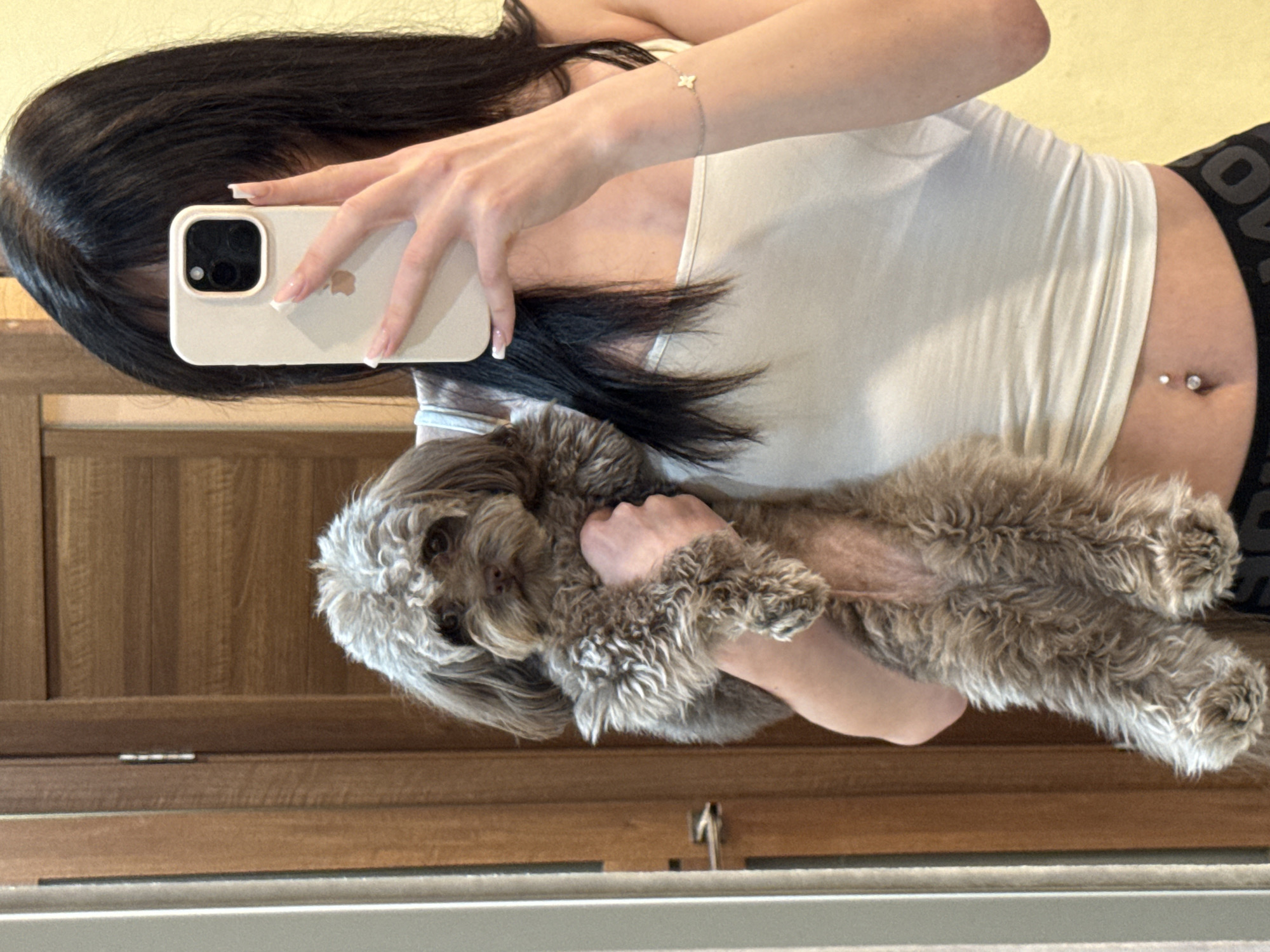 A young woman with long black hair is taking a selfie in a room, holding a small, fluffy brown dog. She is wearing a white tank top and black shorts. A belly button piercing is visible. The background includes a wooden door and a wall. The location is identified as Khimki, Russia.