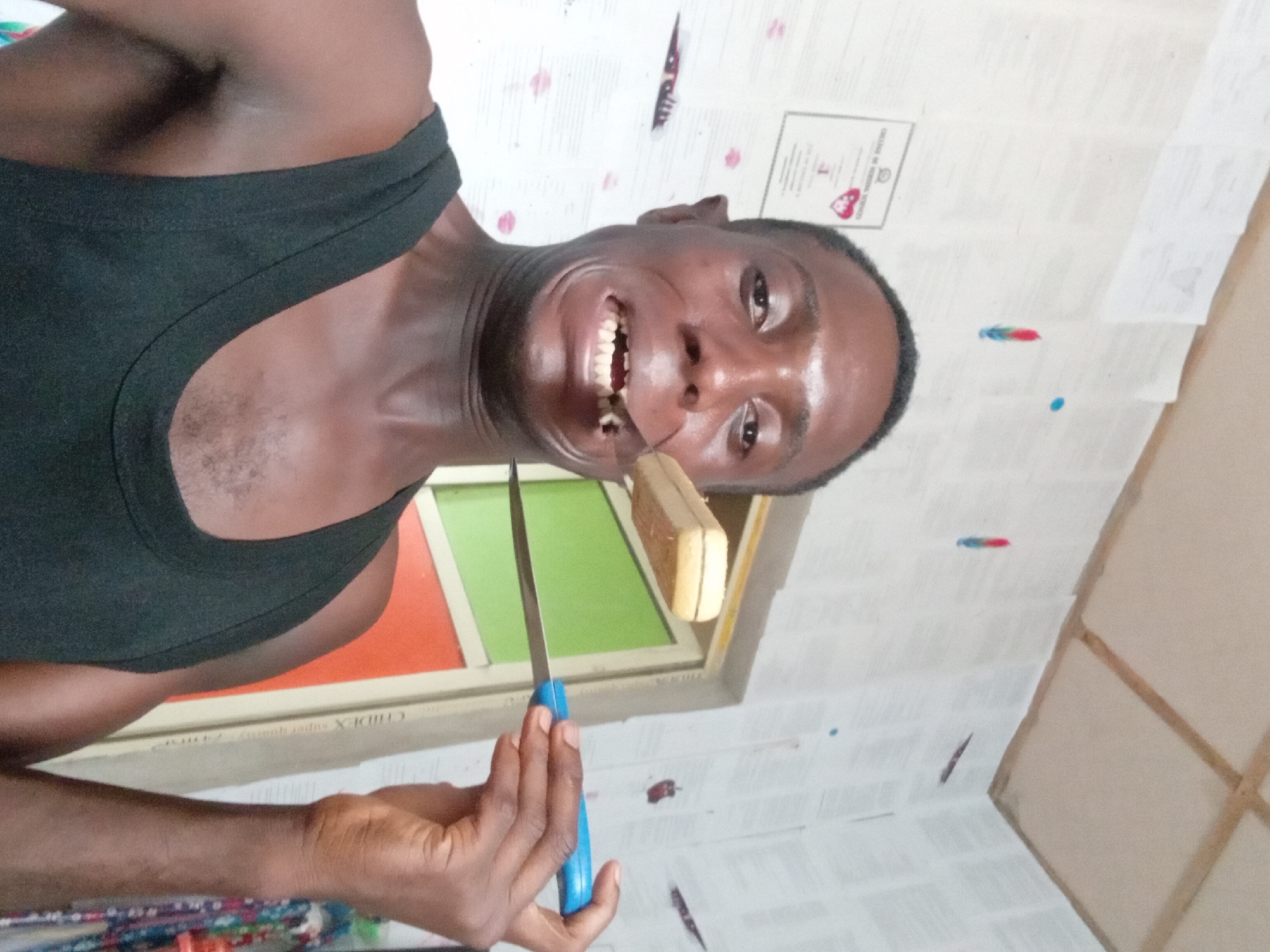 A man with dark skin and a black tank top is holding a knife with a blue handle. He is looking at the camera with his mouth open. He is holding a block of something off-white in his mouth. The background consists of wallpaper covered with what appear to be papers, some with colored marks. In the background, there's a window frame with orange and green segments. The location is possibly Akwete, Nigeria.