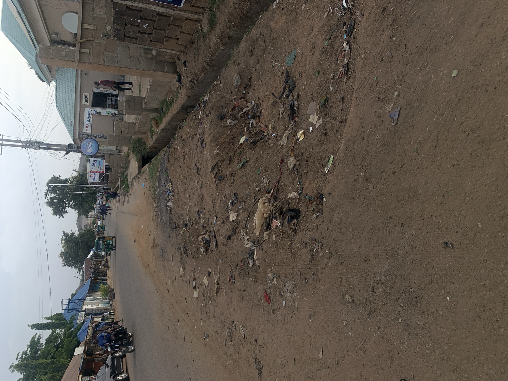 The image shows a dirt road in what is likely New Stadium, Nigeria. The road is littered with trash, including plastic bags, paper, and other debris. A concrete drainage ditch runs along the side of the road, also filled with trash. Buildings are on either side of the road, including a pharmacy, shops, and homes. There are cars, motorcycles, and auto rickshaws on the road. People are walking along the road and interacting with vehicles. The sky is overcast.