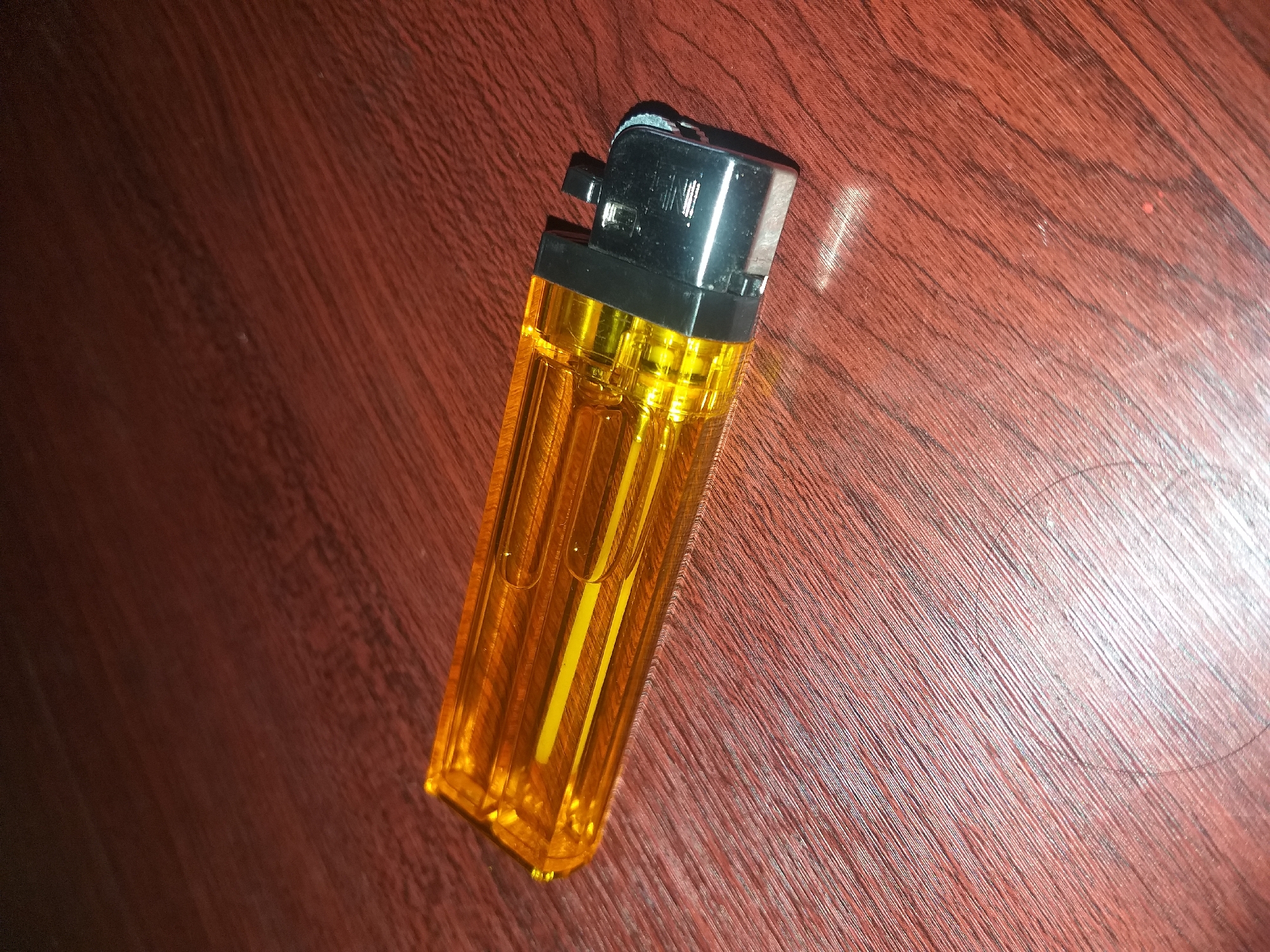 A clear, orange, disposable lighter lies horizontally on a dark brown, wood-grained surface. The lighter is oriented from left to right, with its flint and metal striker mechanism at the right end. The surface appears to be a tabletop or desk. There is no indication of the city of Air Jamban, Indonesia, or any other specific location in the provided image. The lighting in the image suggests an indoor setting. There are no people, actions, or interactions depicted.