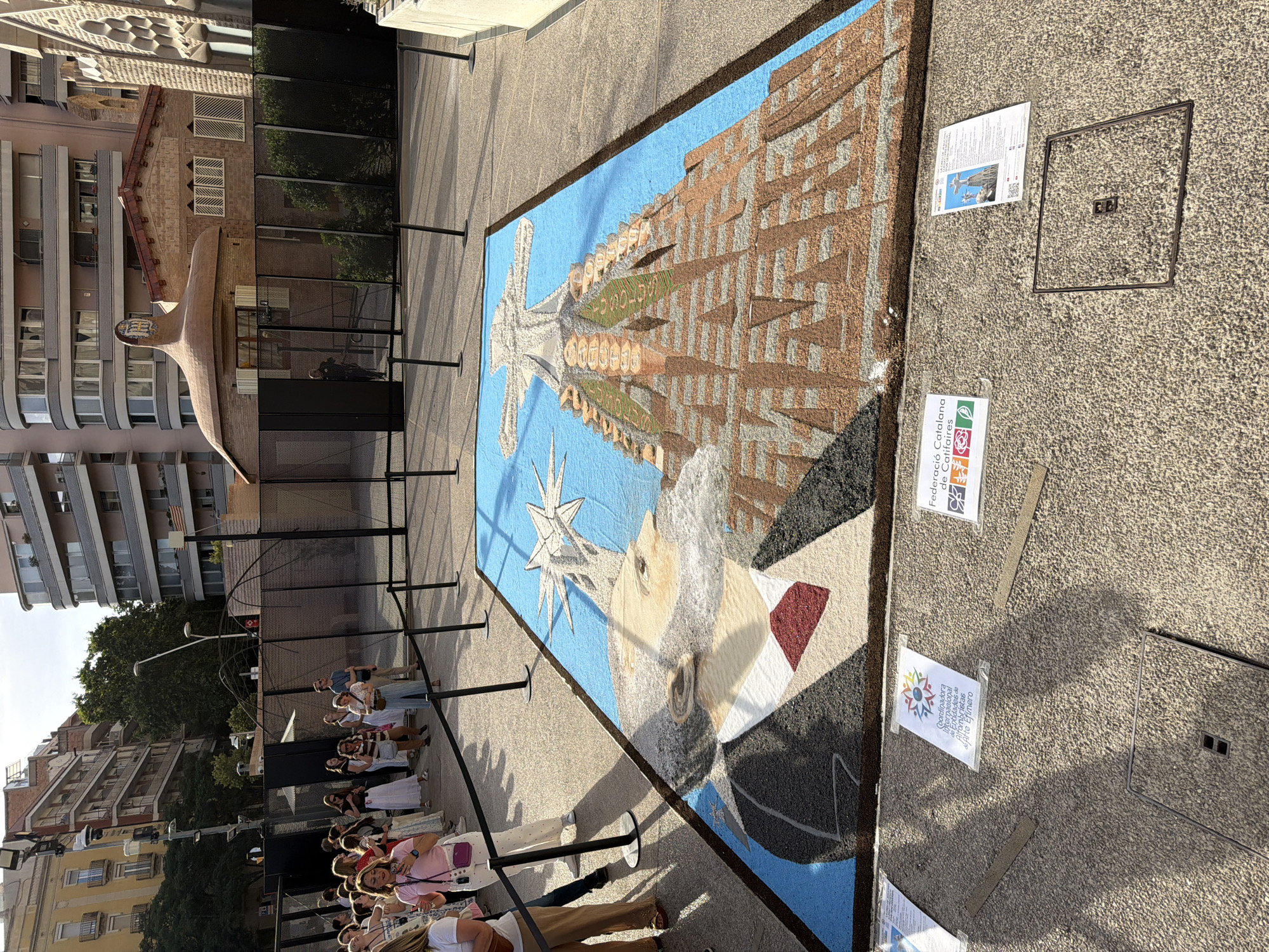 In Barcelona, Spain, an elaborate alfombra de flores (flower carpet) depicts a portrait of Antoni Gaudí, the famed architect, superimposed on a rendering of the Sagrada Familia. The carpet is laid out on a paved surface, bordered by black stanchions and ropes. Several people, likely tourists and locals, are visible in the background, observing the artwork. The Sagrada Familia's distinctive spires and facades are recognizable in the carpet's design, rendered in colorful petals or similar organic materials. The portrait of Gaudí features his characteristic beard and attire, including a suit and tie. Two informational signs with text and graphics are placed on the ground near the carpet. The overall scene suggests an outdoor exhibition or cultural event celebrating Gaudí's legacy.