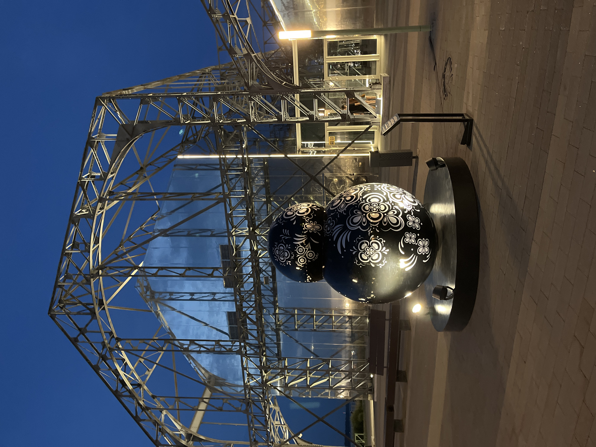 The image depicts an outdoor nighttime scene in Nizhny Novgorod, Russia. In the foreground, a large, black spherical sculpture adorned with intricate white floral patterns is prominently displayed on a circular black base. The sculpture is illuminated by spotlights positioned around its base, casting soft light on the patterned surface. Behind the sculpture stands a large, skeletal metal structure, possibly part of a building or exhibition space, with glass panels reflecting the surrounding lights and the deep blue twilight sky. The ground is paved with brickwork, and several wooden benches are visible in the middle ground. A modern light fixture stands to the right of the sculpture, casting a warm glow. In the background, two individuals, a man and a woman, are seen near the entrance of the metal-framed structure. The overall atmosphere is calm and illuminated by artificial lighting against the darkening sky.