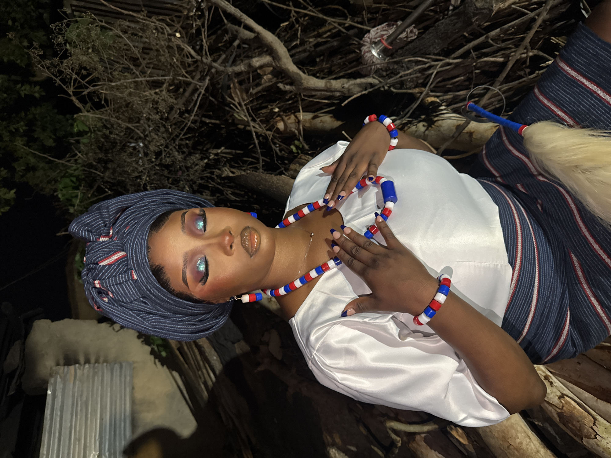 In Gura Top, Nigeria, a person is depicted in a close-up portrait, lying down amidst natural debris and a rustic setting. The subject is adorned in a white satin top and a blue and white striped fabric, possibly a skirt or wrap, with red accents. They wear striking blue and red beaded jewelry, including a necklace and bracelets, and a matching headwrap. The person's makeup is dramatic, featuring iridescent blue eyeshadow and glossy lips. Their nails are painted a deep navy blue. To the right of the subject, a decorative whisk or brush with a light-colored mane is visible, attached to a blue rod. The background consists of dry branches and foliage, suggesting an outdoor environment. The lighting indicates it is nighttime, with a soft illumination highlighting the subject against the darker surroundings. The overall mood is one of serene beauty and cultural expression.