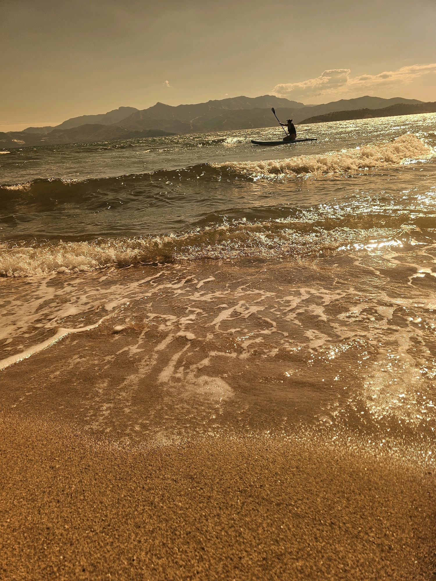 A person is paddleboarding on a body of water with a mountain range in the background. The water is choppy and reflects the sunlight, creating a shimmering effect. Waves are breaking on a sandy beach in the foreground. The sky is hazy and a warm, golden color.