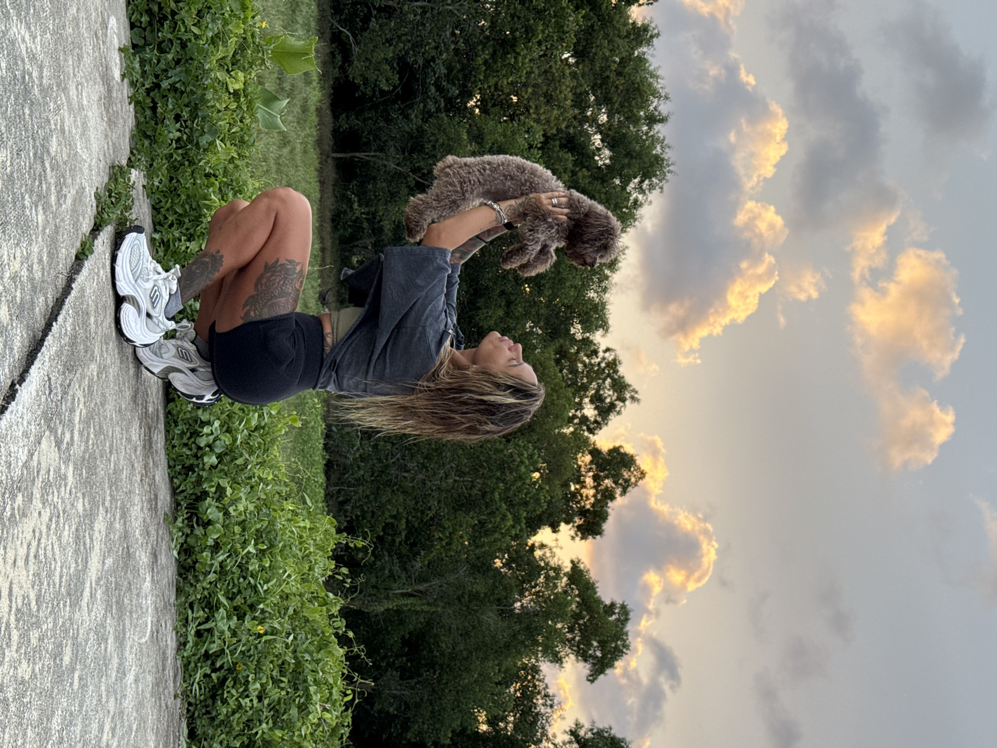 A woman is squatting outdoors, holding a small, brown, curly-haired dog in her arms. The dog is being held up towards the sky, and the woman is looking up at it. The woman has long, blonde, wavy hair, and is wearing a dark grey t-shirt, black shorts, and white athletic shoes. She has tattoos on her legs. The background consists of lush green trees and a cloudy sky with hints of sunset colors. The ground in the foreground is a paved surface with cracks, surrounded by green vegetation. The scene appears to be in a natural or semi-natural outdoor environment.
