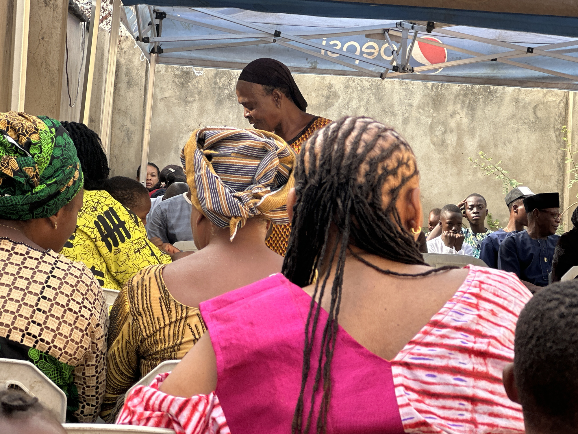 The image captures an outdoor gathering in what appears to be a courtyard or open area, possibly in the city of Unknown, Unknown. A group of people are seated in chairs, facing the same direction. Several women wearing colorful, patterned head wraps and clothing are in the foreground, obscuring the view of others. Behind them, a woman stands, wearing a dark head covering and patterned dress. Further back, a group of seated men and children can be seen. Overhead, a blue and white canopy, bearing the logo of Pepsi, provides shade. The background consists of a gray wall and sparse greenery.