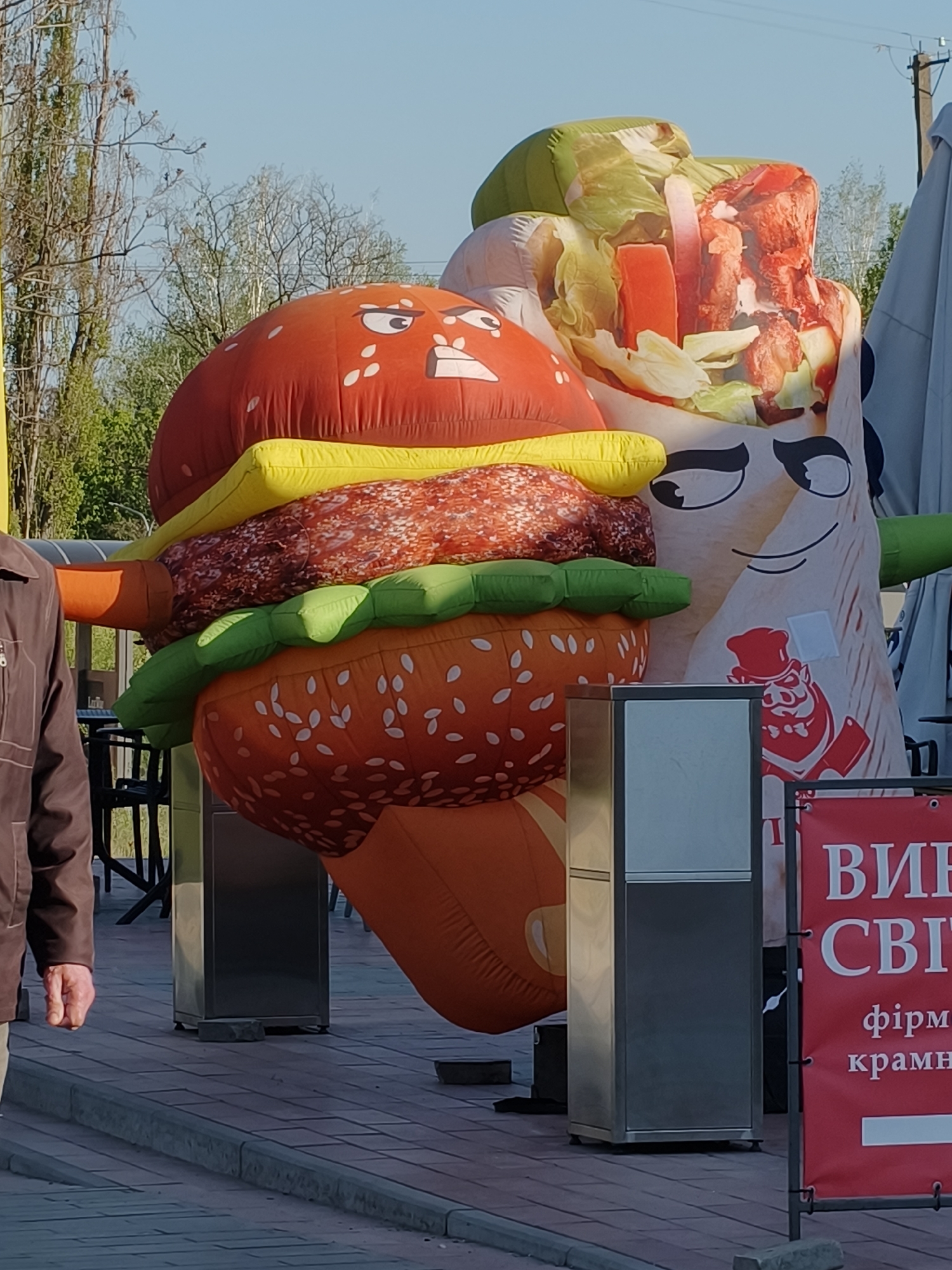 Outside a business in Kryvyi Rih, Ukraine, stands an advertising installation featuring an inflatable burger with an angry face and an inflatable shawarma with a smiling face. The inflatables are positioned on silver metal pedestals. A man in a brown jacket walks by on the left. A sign in Ukrainian is visible on the right. Outdoor seating and trees are in the background.