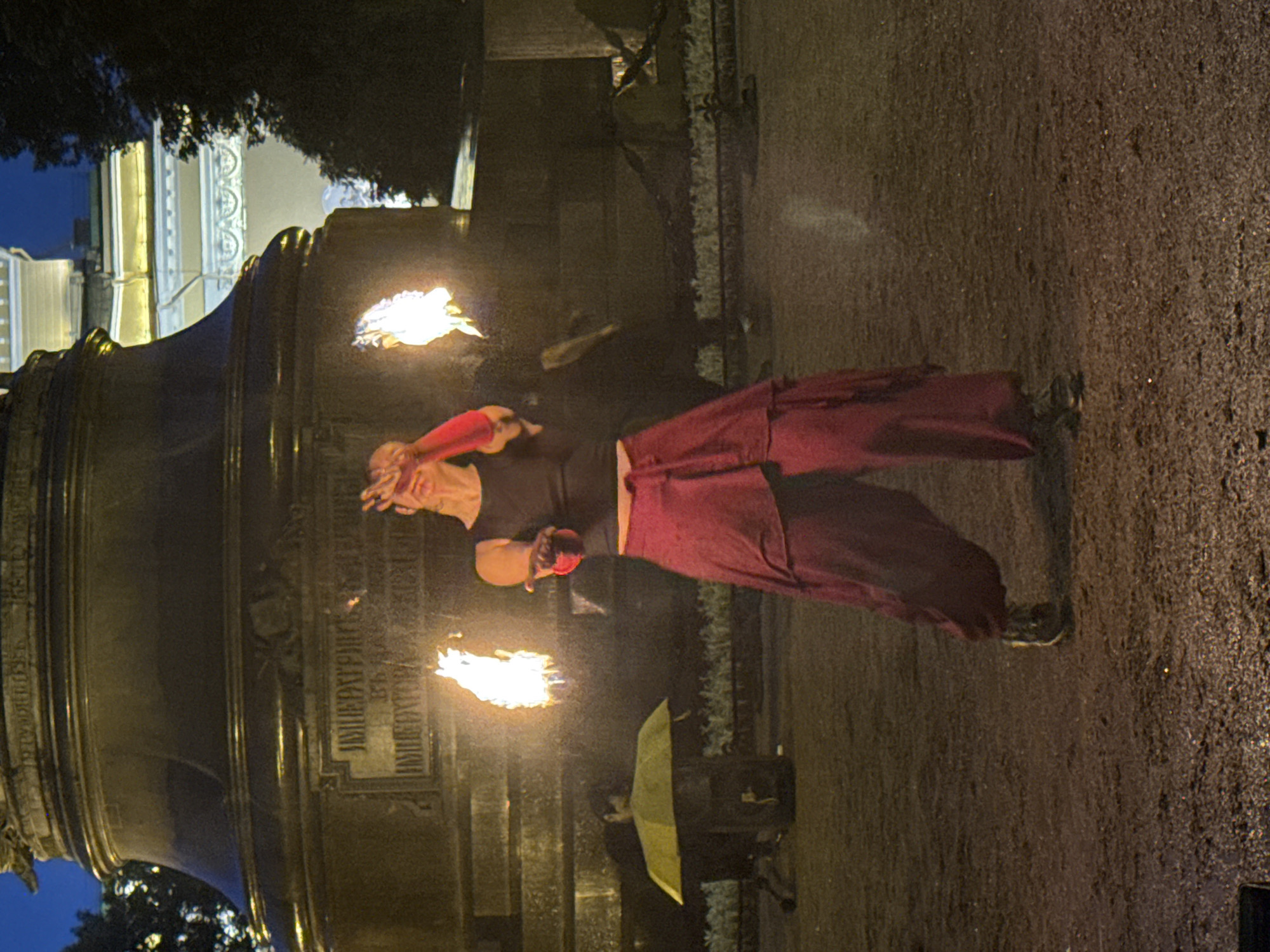 A person is performing with fire in front of a large monument at night in Palace Square, Saint Petersburg, Russia. The person, wearing a dark sleeveless top and voluminous red pants, holds fire in both hands, creating bright flames. The monument, likely the Alexander Column, is dark and imposing, with some inscriptions visible. In the background, a building with illuminated windows is partially visible, and trees frame the scene. A yellow umbrella is set up on a stand to the left of the frame, casting a shadow. The ground is covered in gravel or dirt. The overall lighting suggests it is dusk or night, with artificial light illuminating the performance and the building in the background.