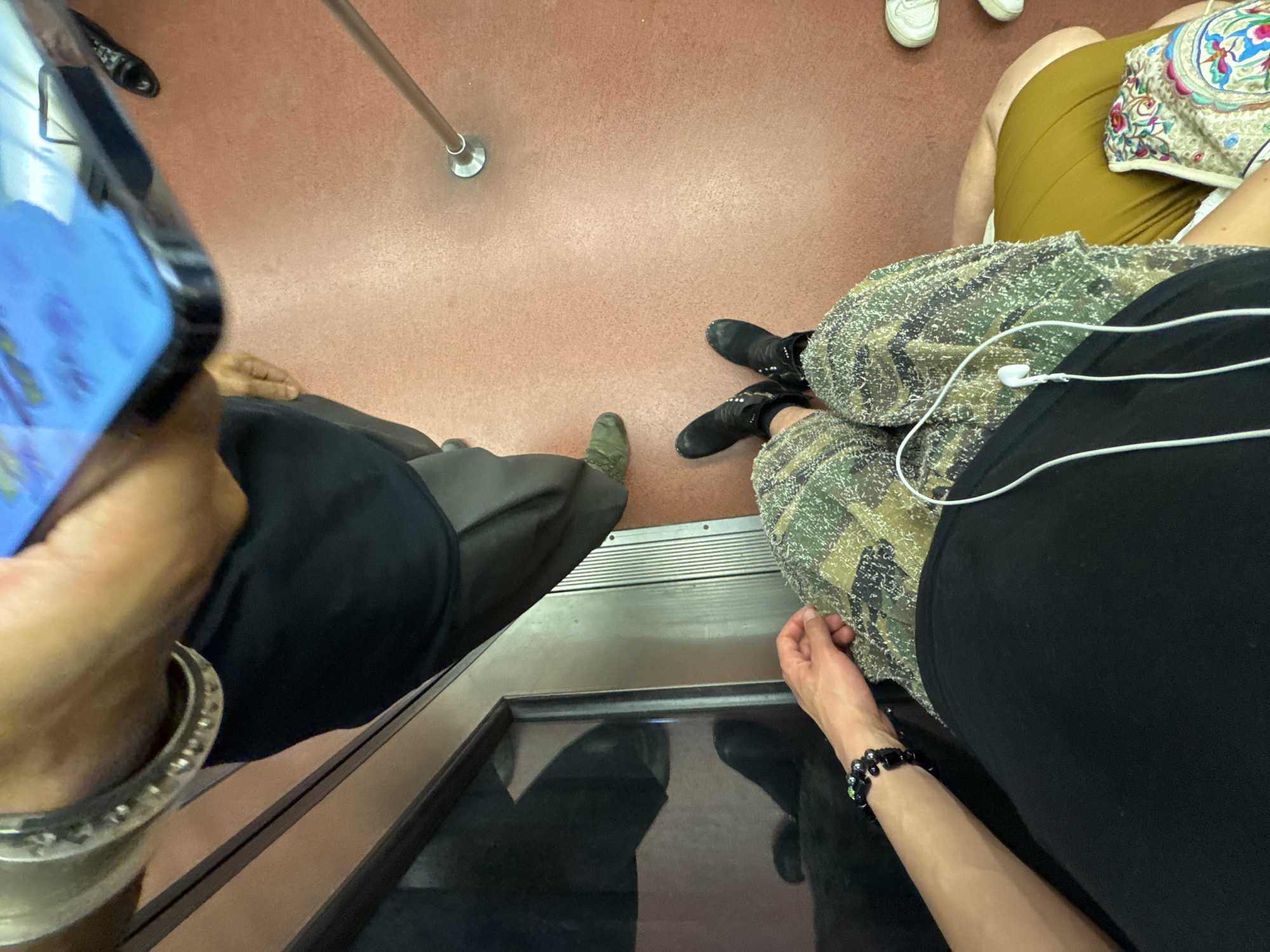 The image captures a view from above, looking down into what appears to be the interior of a Paris Metro train. The floor is a reddish-brown color. A silver pole runs vertically. Three sets of feet and legs are visible. The viewer sees a person's hand holding a phone, with a large silver bracelet visible on their wrist. The person is wearing a black shirt and dark pants, and one green sneaker is also visible. Another person is wearing a patterned fabric with camouflage print, black boots, and a black shirt. This person also has a bracelet on their arm. A third person sits in a yellow skirt and has a patterned bag on their lap, and white shoes are visible. The shot is taken from an elevated position, looking down towards the floor of the train.