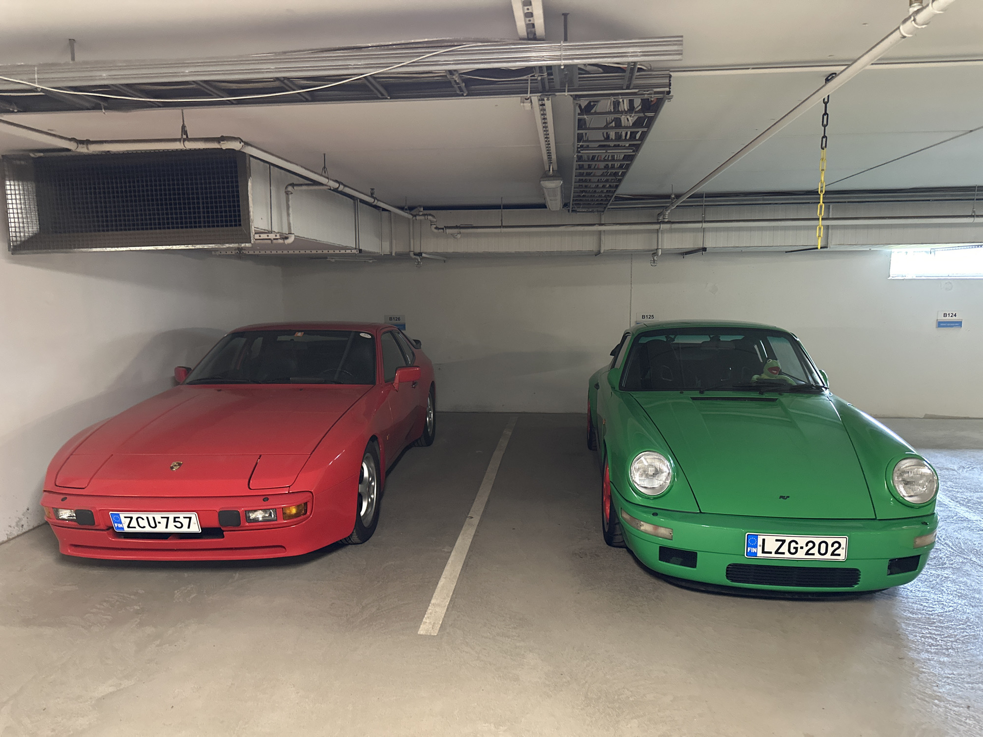 The image depicts a brightly lit underground parking garage in Espoo, Finland. Two classic Porsche sports cars are parked side-by-side. On the left is a vibrant red Porsche 944 with the license plate ZCU-757. To its right is a striking green Porsche 911 Carrera 2 (964 generation) with the Finnish license plate LZG-202. The cars are parked in designated spots marked by white lines on the concrete floor. Above the vehicles, the ceiling features extensive electrical conduits, ventilation systems, and lighting fixtures. A chain with yellow and black links hangs from the ceiling near the green Porsche. The overall environment is clean and well-maintained, characteristic of a modern parking facility.