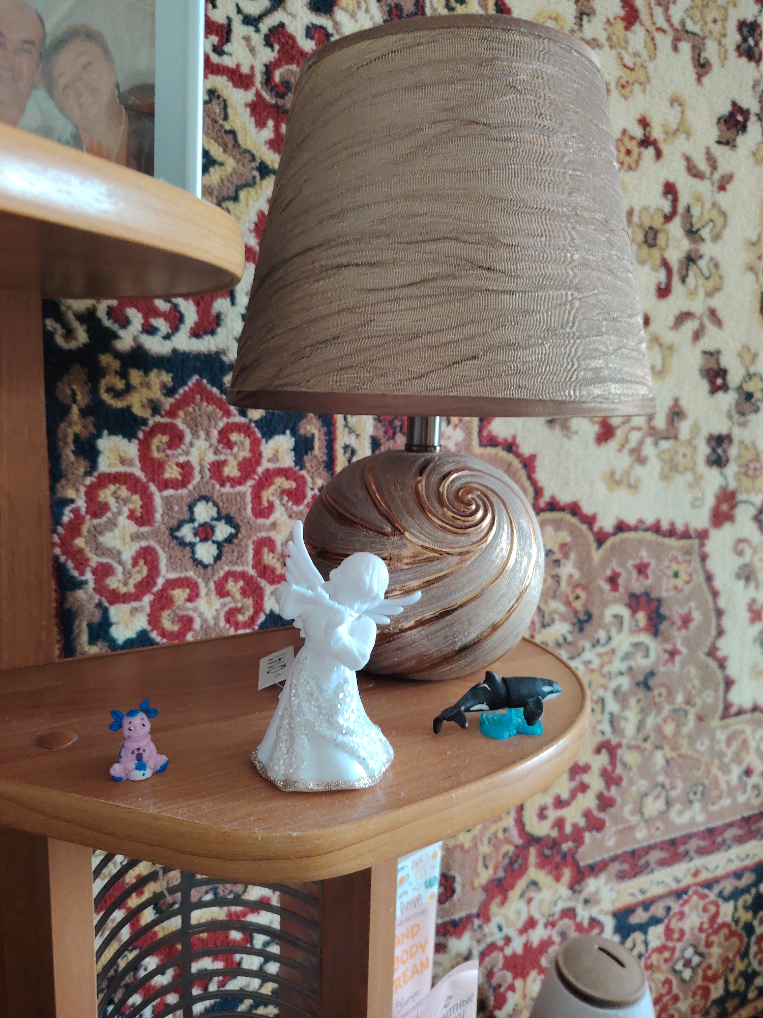 The image depicts a close-up view of a wooden shelf with various decorative items. On the left, a small, light pink figurine with blue accents, resembling a cartoon character, sits on the shelf. To its right is a white angelic figurine with glittery accents on its dress, holding what appears to be a musical instrument. Further to the right, a miniature black and white orca toy rests on a small blue platform. Behind these figurines, a large lamp with a textured, golden-brown shade stands prominently. The lamp base is a decorative, swirling design in beige and gold tones. The shelf itself is made of light-colored wood and has a rounded edge. The background is a richly patterned carpet with floral motifs in red, beige, blue, and black. A framed photograph of two smiling individuals is visible in the upper left corner, partially obscured by the shelf. The overall scene appears to be an indoor setting, likely a living room or display area. There is no discernible information within the image that can place it in Rostov-on-Don, Russia.