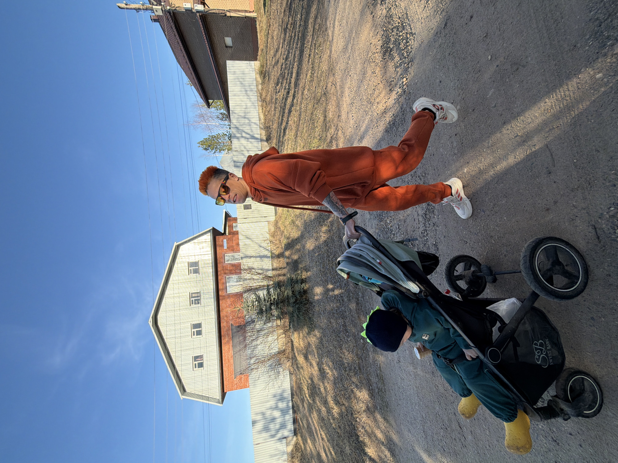 The image depicts an adult pushing a child in a stroller on an unpaved path in Chaikovsky, Russia, under a clear blue sky. The adult, positioned on the left, is wearing an orange hooded tracksuit, white sneakers with red accents, and sunglasses. They have short red-orange hair and tattoos visible on their left arm. They are looking towards the camera with a slight smile.
The black stroller, bearing the visible text "SWEET BABY" and "S&B," contains a child dressed in a dark green hooded tracksuit, bright yellow boots, and a dark beanie with distinctive green spikes. The child's face is not visible. A small white emblem is present on the child's tracksuit.
The background features several buildings: a brick house with a corrugated metal roof and a larger, multi-story building with a white corrugated metal facade and roof. A corrugated metal fence runs parallel to the path. Sparse trees and dry grass are visible behind the fence and buildings. A utility pole with power lines extends across the clear sky. Strong shadows are cast on the ground, suggesting bright sunlight during the day. The path itself is made of dirt and gravel.