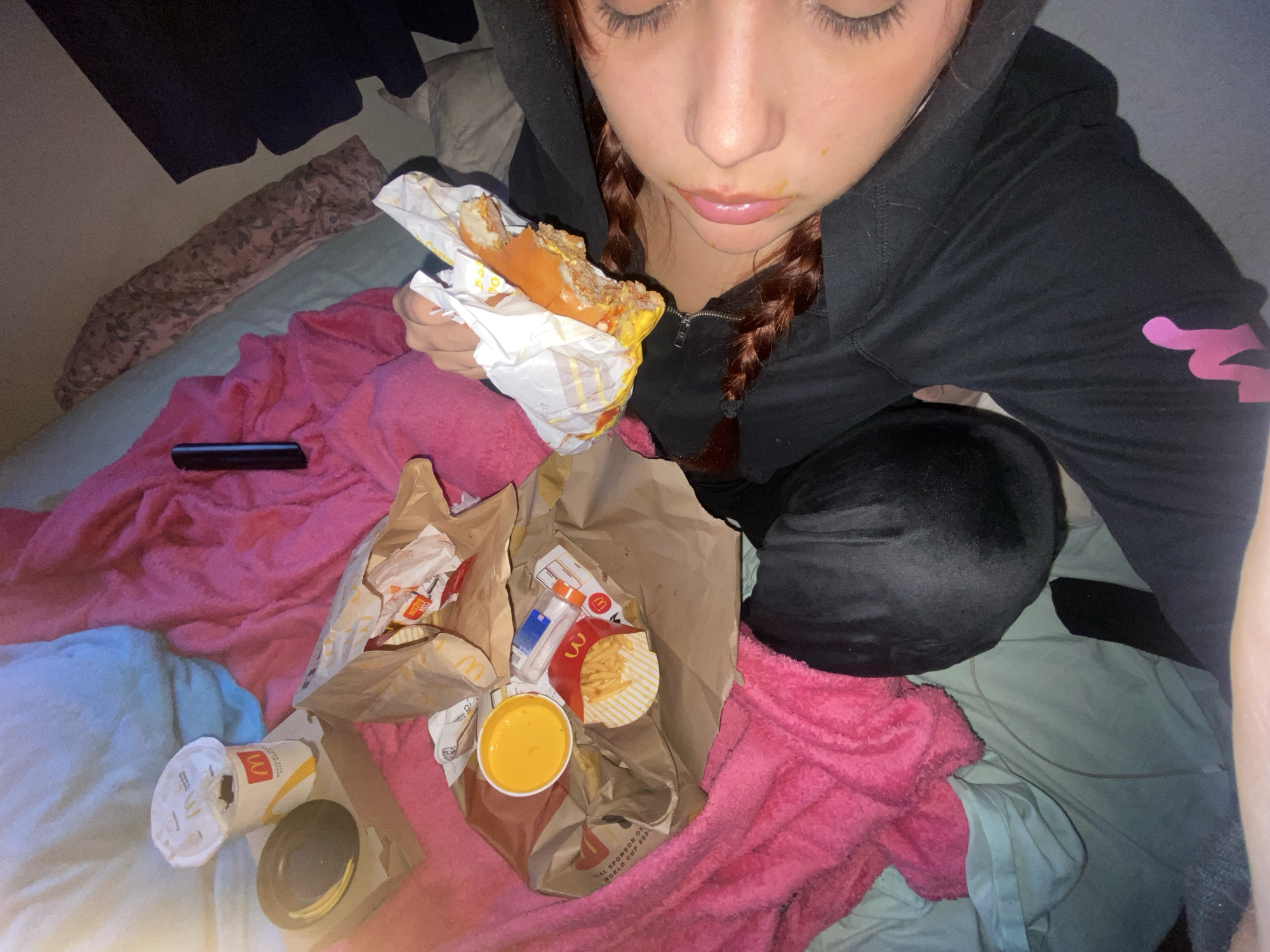 A person with reddish-brown hair in braids is seated on a bed with light blue sheets. They wear a black hoodie and black sweatpants. The person is holding a half-eaten McDonald's burger wrapped in paper. Around them on the bed, are two McDonald's paper bags, one containing fries and a container of sauce, and another with burger wrappers and condiment packets. Also on the bed are a McDonald's cup and a black object. There is also a pink and light blue blanket. The location is not discernible from the image.