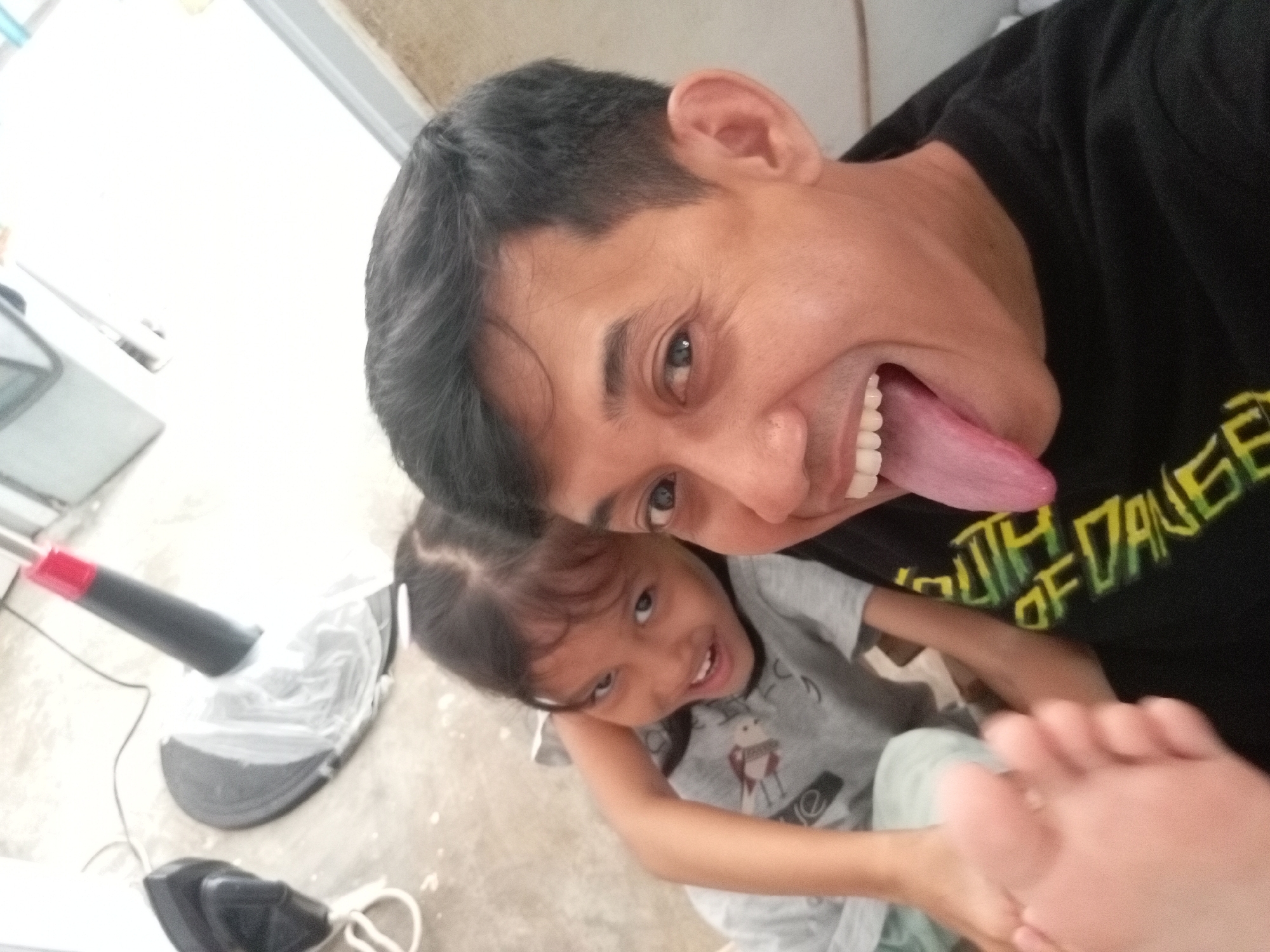 An over-the-shoulder shot shows a man with his tongue sticking out and a young girl looking at the camera, in what appears to be an indoor setting in Batam, Indonesia. The man is wearing a black t-shirt with yellow text that reads "SOUTH OF DANGER". The girl is wearing a grey t-shirt with a cartoon character. The floor appears to be concrete and there is a white plastic-covered object and a vacuum cleaner in the background.