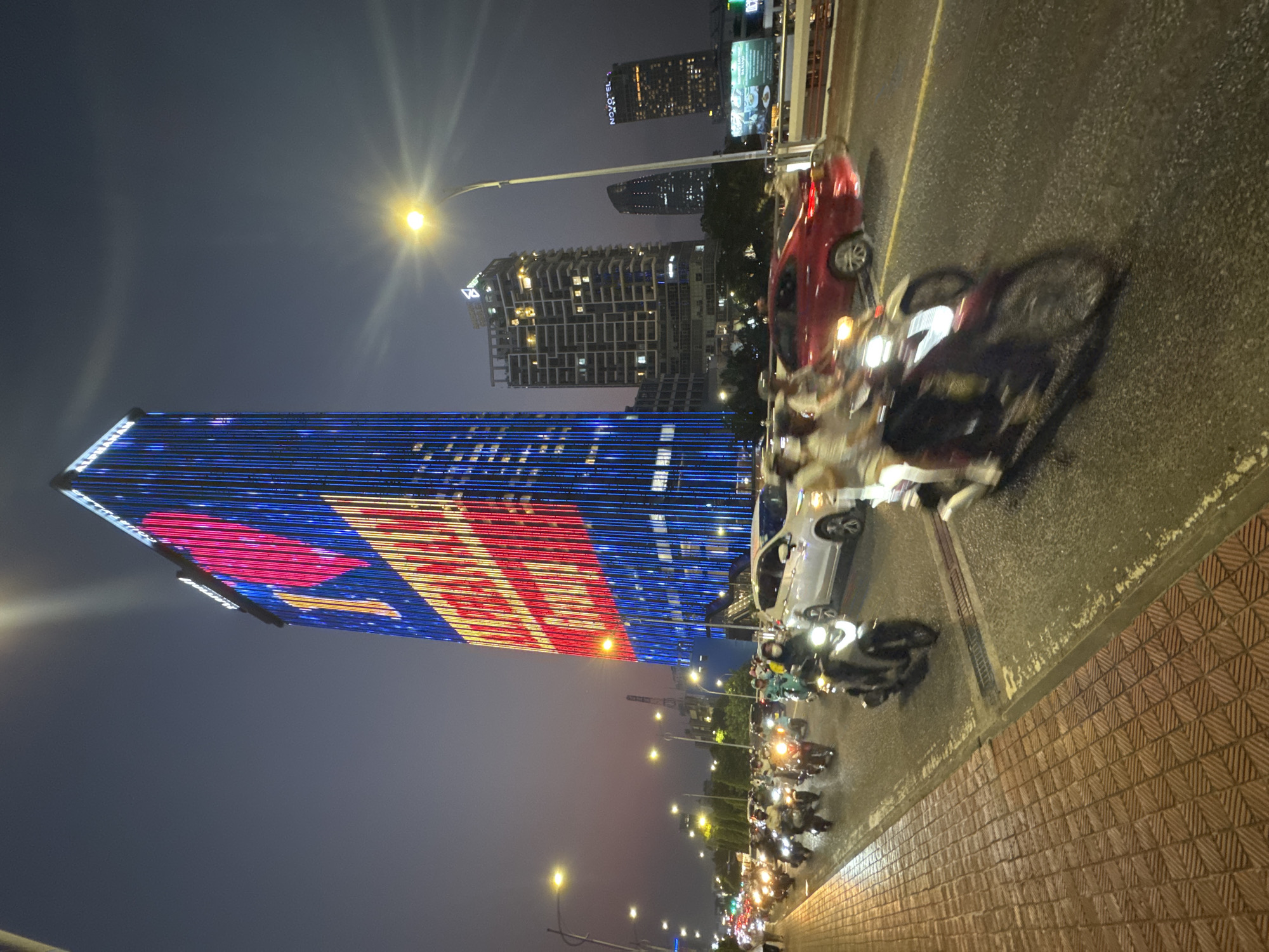 The image shows a night scene in Đà Nẵng, Vietnam. The Alpha Asia Tower stands tall with colorful lights, featuring red and blue stripes. The street below is filled with numerous scooters and a few cars moving along the road. The sky is dark, illuminated by streetlights and building lights. The pavement is bricked and appears wet.
