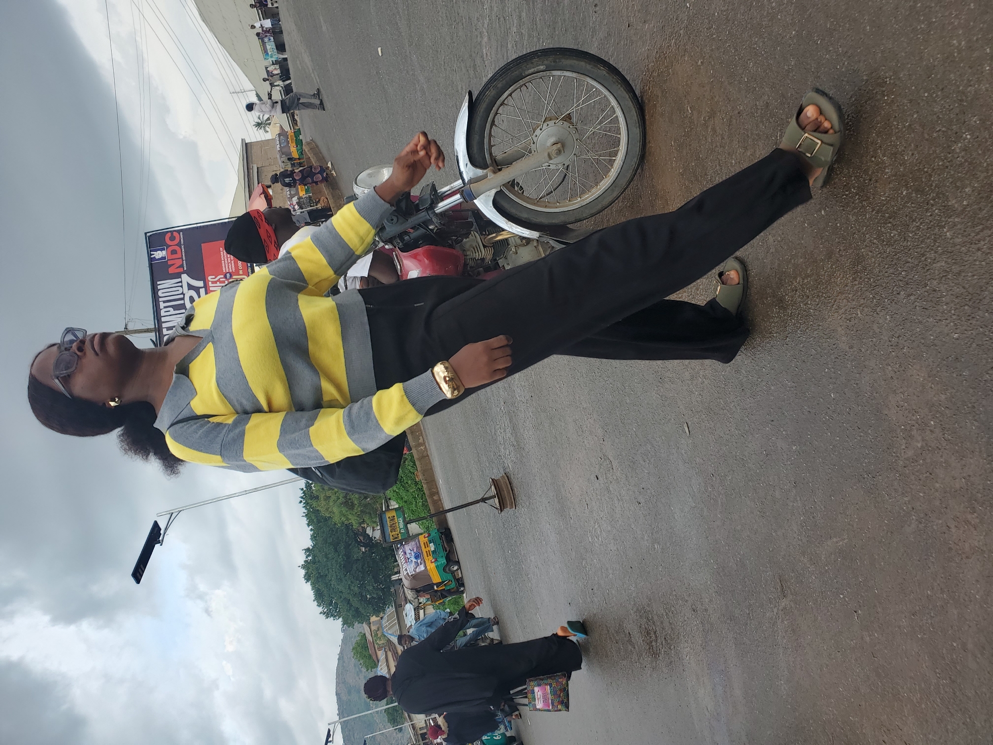 A medium shot captures a woman walking towards the right of the frame on a wet asphalt road. She wears a horizontally striped yellow and gray long-sleeved shirt, black trousers, and olive-green sandals. A gold bracelet adorns her left wrist. To her right, a red motorcycle is parked, and a portion of a man wearing a red and black patterned headscarf is visible on it. In the background, several other people are walking, and a yellow and green auto-rickshaw is seen. A large banner with "REDEMPTION 2027 NDC" and other text is displayed behind the people. The environment appears to be an urban street in Gwong, Nigeria, with a cloudy sky overhead. The road is wet, suggesting recent rain. There are trees and buildings in the distance, typical of a city scene.