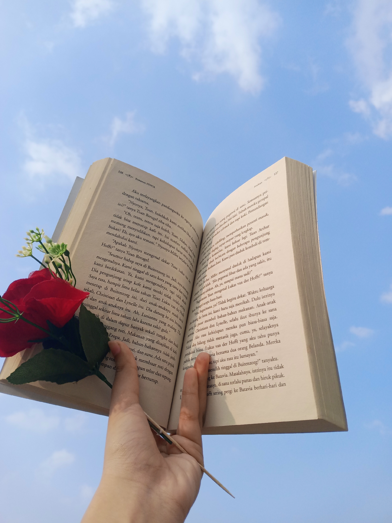 A person's hand holds an open book against a bright blue sky with scattered white clouds. The book is positioned at an angle, with the left page facing upwards and the right page downwards. A red artificial rose with green leaves is placed on the left page, near the spine of the book. The text on the pages is in Indonesian. The image is taken from a low angle, looking up at the book and the sky. No specific location in the city of Demak, Indonesia, is depicted or suggested by the image.