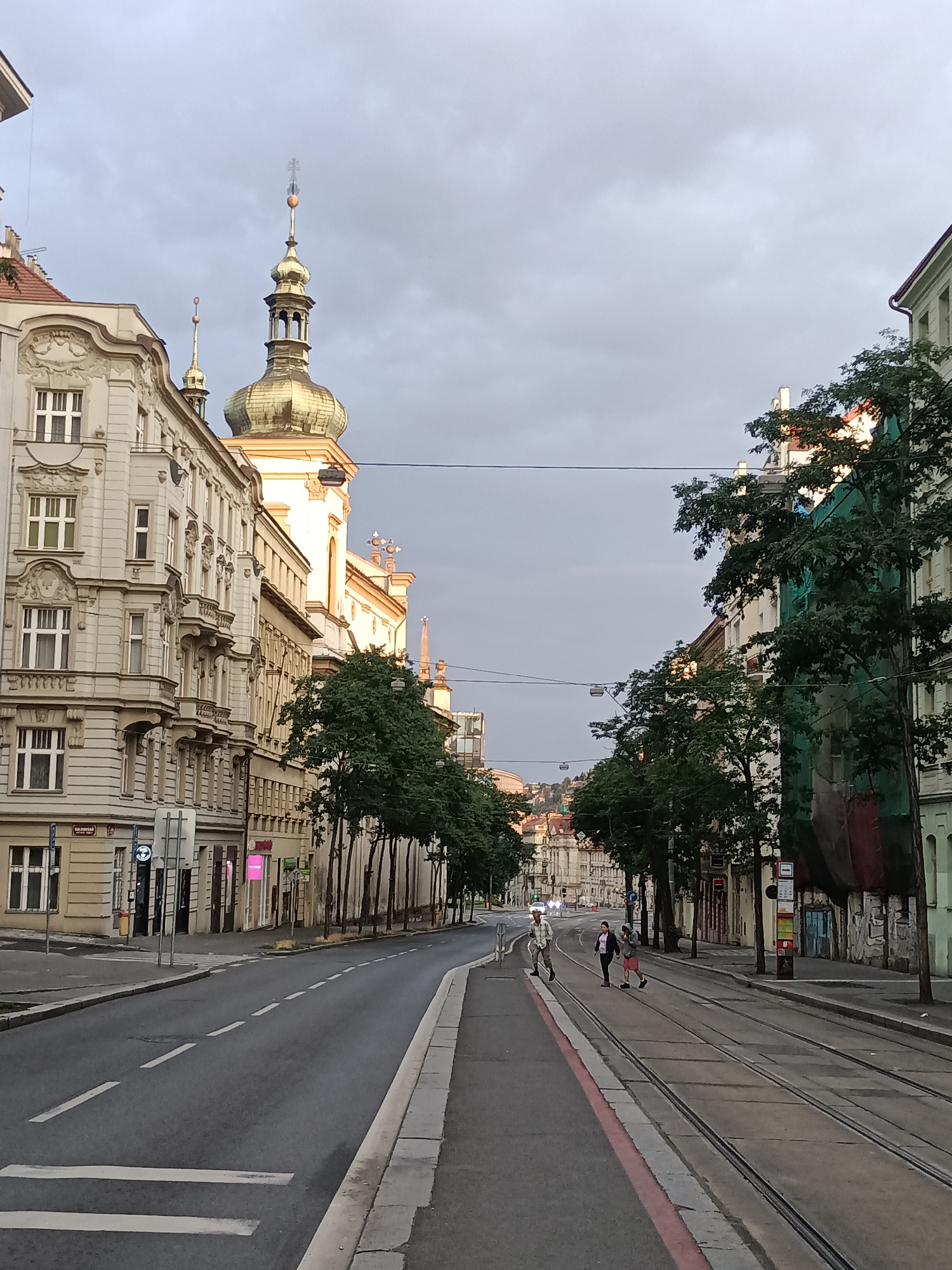 The image depicts a street scene in Prague, Czechia, likely in the Old Town or Lesser Town, characterized by its historical architecture and tram infrastructure. The perspective is from street level, looking down a road lined with ornate buildings. On the left, a grand, light-colored building with intricate carvings, balconies, and numerous windows dominates the foreground. Adjacent to it, a church or religious building with a prominent golden dome and spire stands tall, catching the soft light of what appears to be either early morning or late afternoon. The sky is overcast with a heavy, gray cloud cover.
On the right side of the street, a line of trees with dark green foliage partially obscures the facades of buildings, some of which appear to be under renovation, indicated by green netting draped over a section of one building. Tram tracks run parallel to the sidewalk on the right.
In the middle ground, three individuals are crossing the street at a pedestrian crossing. One person, on the left, appears to be wearing a light-colored shirt and dark pants, possibly a man. To their right, a woman in a dark jacket and pink shorts is walking, followed by another person in a grey hoodie and red shorts. The street itself is paved, with distinct lane markings and a pedestrian crossing marked with white stripes. The overall atmosphere is subdued, likely due to the cloudy weather. The presence of tram lines and the architectural style strongly suggest a European city, and given the specific architectural details like the domed spire, it is highly indicative of Prague.