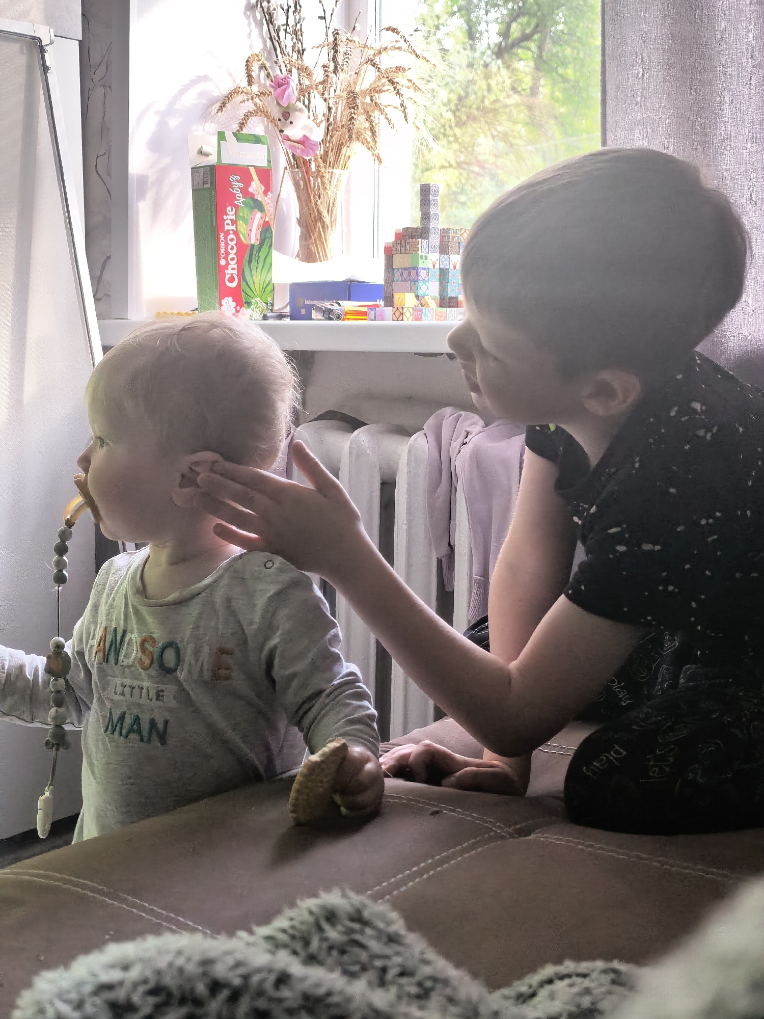 In a sunlit room, two young boys are seated on a brown, textured sofa. The boy on the left, a toddler with fair hair, is wearing a gray long-sleeved shirt with the words "HANDSOME LITTLE MAN" printed on it. He has a pacifier in his mouth, attached to a string of beads and a white toy. His right ear is being gently touched by the hand of the boy on the right. This older boy, with dark hair, is wearing a black t-shirt with a speckled pattern and dark shorts. He is leaning forward, resting his chin on his hand, and looking at the toddler.
On a windowsill behind the boys, a variety of items are visible. There is a carton of "Choco-Pie" juice with a watermelon design, a glass vase filled with dried wheat stalks and a small pink bunny toy, and a stack of colorful building blocks. A radiator is positioned against the wall, with a light purple garment draped over it. Outside the window, greenery and trees are visible, suggesting an outdoor environment beyond the room. The room appears to be a domestic setting, possibly a living room. The image was taken indoors, with natural light streaming in from the window. The scene captures a moment of sibling interaction.