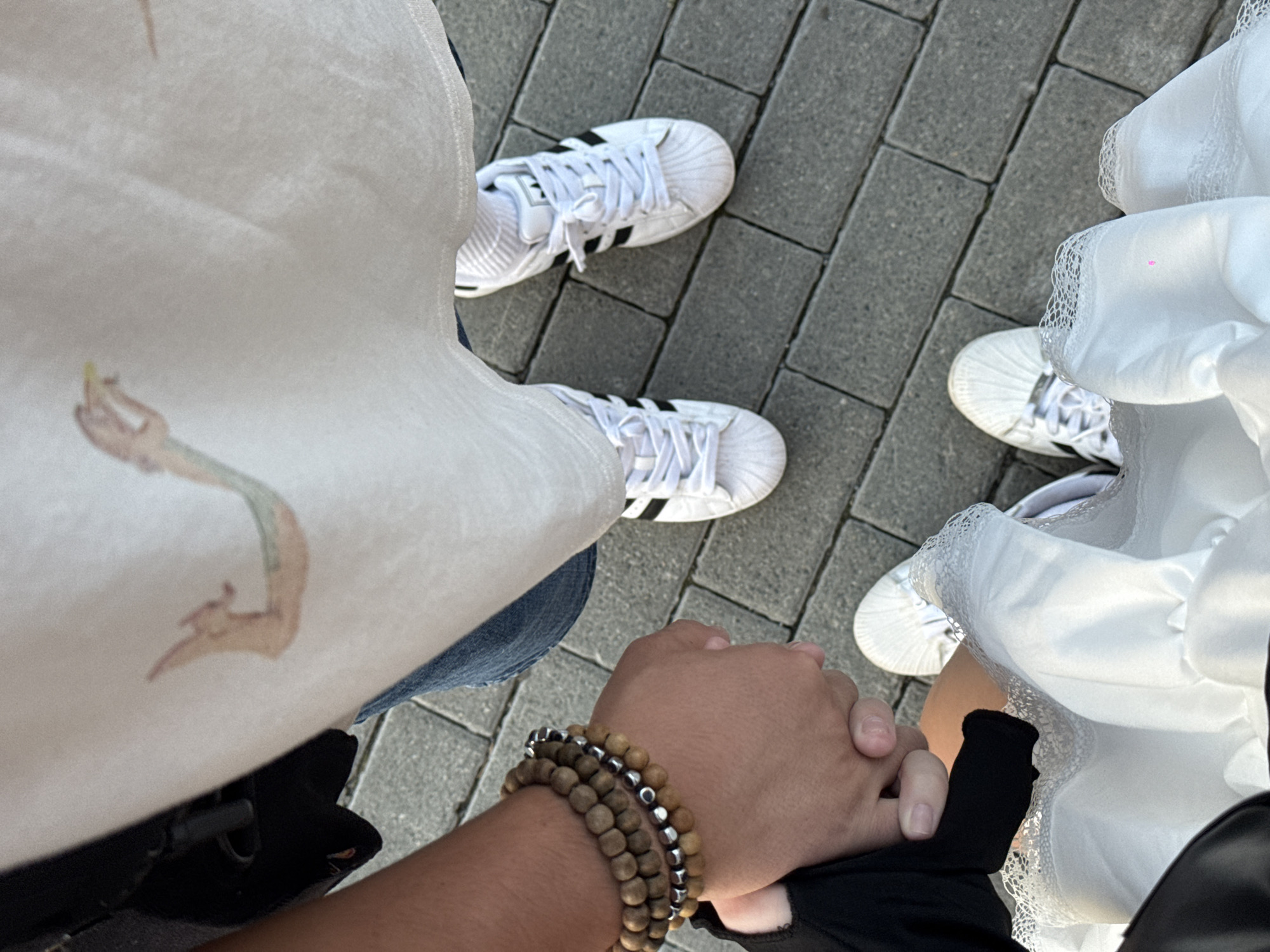 The image shows a top-down view of two people holding hands while standing on a paved sidewalk. The person on the left is wearing a black sleeve and has a bracelet made of wooden beads and silver accents on their wrist. The person on the right is wearing blue jeans and a white t-shirt with a graphic design of a dragon. They are both wearing white Adidas sneakers. The pavement is made of grey rectangular bricks. The image does not provide any information to determine the location in the city of Chelyabinsk, Russia.