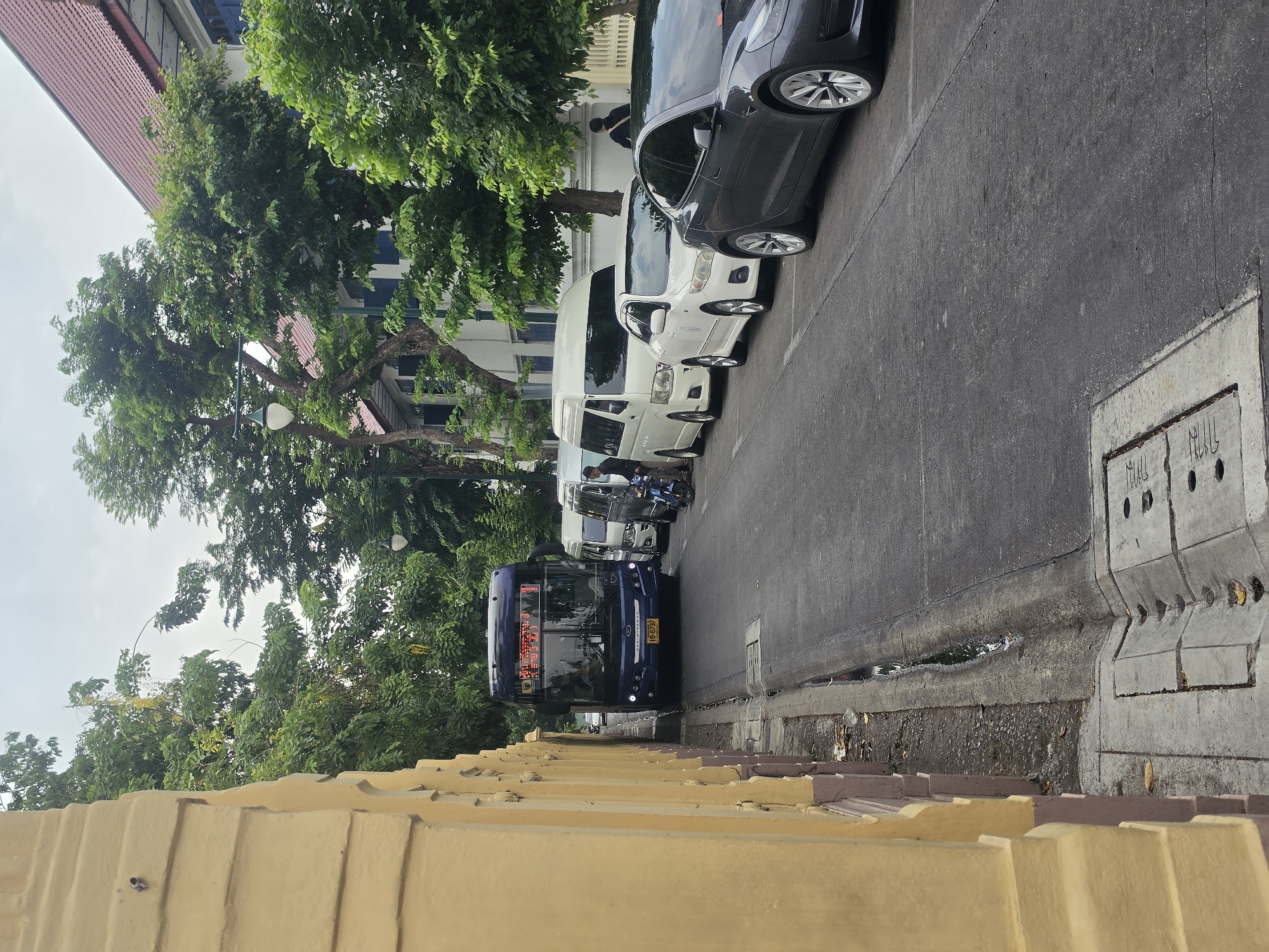 The image depicts a street in Bangkok, Thailand, showing a bus moving down the road with several parked cars and vans along one side. Lush trees line the street, and a yellow wall is visible in the foreground. The street is paved and shows some signs of wear. A sidewalk with a drainage system is visible on one side. The weather appears overcast.