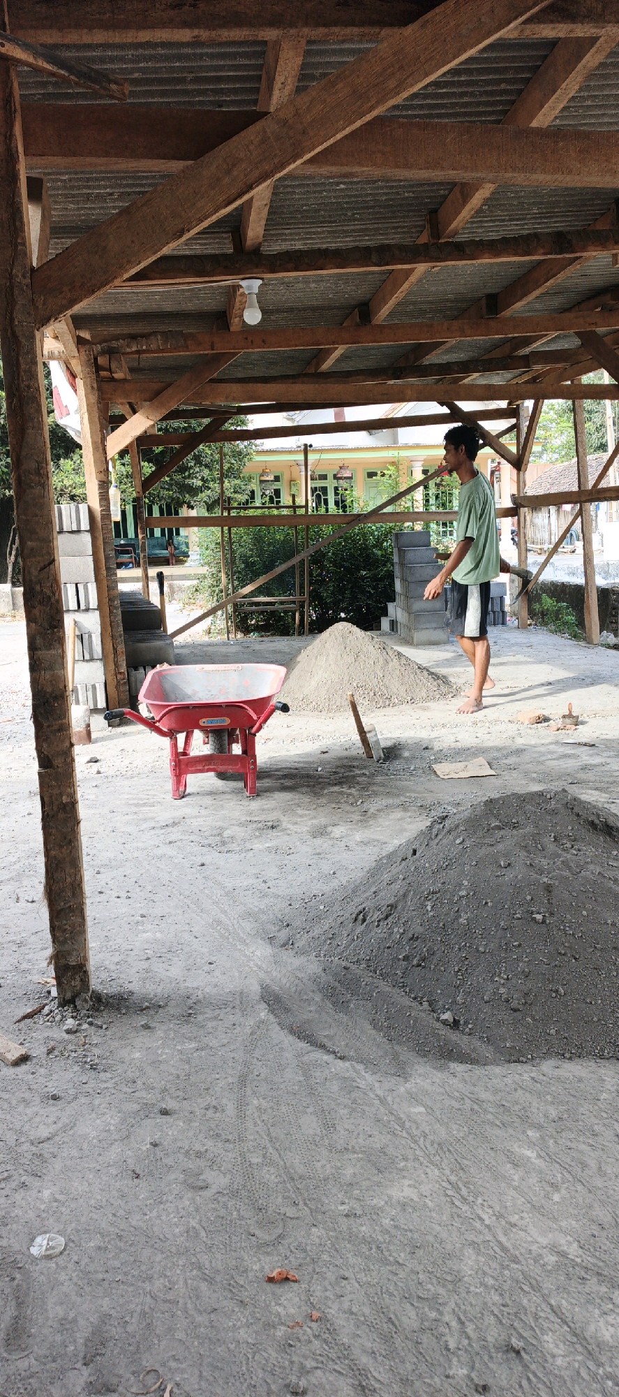 An outdoor construction site in Kasiyan, Indonesia, features a young man wearing a green t-shirt and dark shorts, walking barefoot. He is near a pile of grey sand. In the foreground, a red wheelbarrow is positioned on the sandy ground. The construction area is partially covered by a wooden-framed roof with a corrugated metal sheet ceiling, a light bulb hanging from it. In the background, a building with green window frames and doors is visible, surrounded by foliage. The ground is a mix of sand and dirt with tire tracks. Stacked concrete blocks are visible on the left side of the frame.