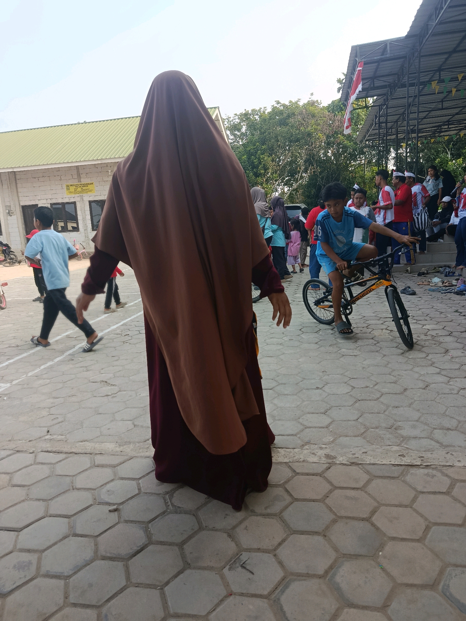 In Swarga Bara, Indonesia, an outdoor event is taking place on a paved, hexagonal-tiled ground. In the foreground, a person wearing a long, brown hijab and a maroon dress stands with their back to the camera, obscuring the view of other participants and the immediate surroundings. Behind them, a young boy in a blue t-shirt and shorts is seated on a black and yellow bicycle, looking towards the camera with a neutral expression. Other individuals, including children and adults, are visible in the midground and background. Some are standing, watching the event, while others appear to be in conversation. A building with a green roof and white walls, possibly a school, is situated on the left side of the image, with a sign that reads "GEDUNG PENDIDIKAN". A covered stage or pavilion with a metal roof stands on the right, adorned with Indonesian flags and triangular green and yellow flags. The sky is clear and bright, suggesting a daytime setting. The overall atmosphere appears to be that of a community gathering or school event.
