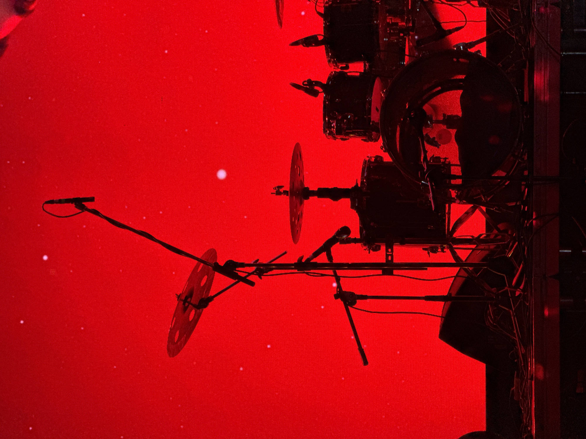 This image captures a drum kit and associated equipment silhouetted against a vibrant, intense red background, likely part of a stage during a live music performance or rehearsal. The scene is dominated by the strong red light, which illuminates the backdrop as if it were a large LED screen or a vividly lit stage curtain.
The main subjects are the various components of a drum kit, including several drums of different sizes and cymbals. These are rendered in dark silhouettes, indicating they are lit from behind or from the sides. Microphones on stands are positioned around the drum kit, suggesting amplification for recording or live sound. A complex web of cables can be seen connecting different pieces of equipment, typical of a professional stage setup. On the far right, parts of what appear to be large speakers or stage elements are also visible.
A notable detail is the faint reflection of a person's silhouette within the surface of one of the larger drum heads, presumably the drummer, adding a human presence to the otherwise inanimate equipment. Small, bright white specks are scattered across the red background, which could be dust particles catching the light, or artifacts from the digital display. The overall mood conveyed is dramatic and energetic, typical of a concert environment. No visible text is present in the image. The event is taking place in Sofia, Bulgaria.