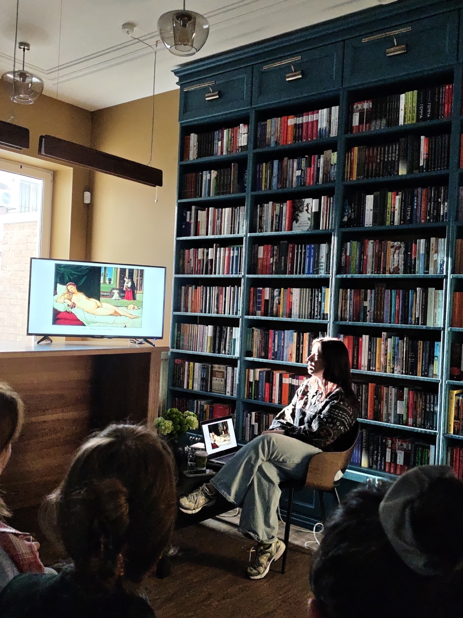 A woman is sitting on a chair, facing left, with her legs crossed in front of a tall, teal-colored bookshelf filled with books. She is wearing a patterned shirt, light blue jeans, and sneakers. The bookshelf spans the height of the wall behind her, and at least four people are seated in front of her, facing the bookshelf. The backs of their heads are visible in the foreground. To the left of the woman, a flat-screen television is displaying an image of a reclining nude figure. On the table near the woman, there is a laptop that also displays the same nude figure. The scene appears to be a presentation or lecture.
Although it is not clear that the location is Vinnytsia, Ukraine without additional information in the image, if it is the case that the location is Vinnytsia, Ukraine then this information could be useful in understanding the cultural context of the scene. The image could be interpreted as showing an educational setting in which the art and culture of Vinnytsia, Ukraine is being shared with an audience.