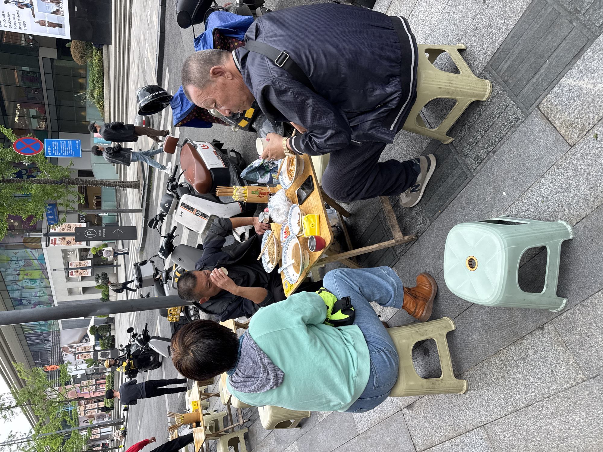 The image captures a street scene in Jinjiang District, Chengdu, China. Two men and a woman are seated on low plastic stools at a small wooden table set up on the sidewalk. They appear to be eating noodles from bowls. The men are wearing dark jackets, while the woman wears a light blue hoodie and jeans. A yellow wrist support is visible on her left hand. The table holds several bowls, chopsticks, and a red can. Parked scooters surround the eating area, including a white scooter with brown accents and black helmets. In the background, pedestrians walk along the sidewalk. There are signs for "Parking" and "SANRI) GIFT GATE", and a "No Parking" sign is visible. The architecture includes modern buildings and trees line the street. The paving is made of gray stone slabs.