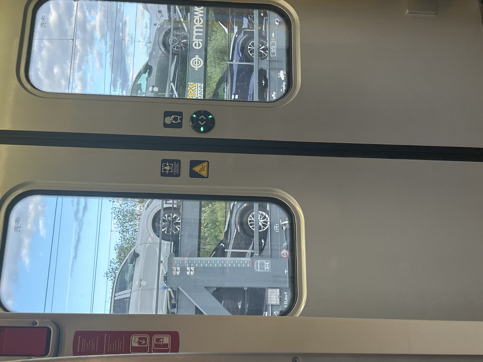 The image, taken from inside a train, shows a view through two rectangular windows with rounded corners. Outside the windows, a car-carrying freight train is visible, suggesting the viewer's train is either moving alongside it or stopped next to it. The freight train carries multiple cars, including dark-colored and white SUVs or crossovers, some with alloy wheels clearly visible. Beyond the freight train, a blue sky with white clouds is seen, indicating daytime. Green foliage is also visible along the trackside.
On the interior panel between the two windows, several controls and symbols are present. A green illuminated circular button with up and down arrows is centered. To its left, a square icon shows a hand pressing a button, possibly indicating a door or window control. Below this, another square icon depicts a box with an inward-pointing arrow, and next to it, a yellow triangular warning sign with an exclamation mark and a falling person symbol, likely indicating caution for slippery surfaces.
Through the upper window, text "Enmew" or "ermew" and a logo resembling a stylized "E" or a circle with a dot are visible on the side of the freight train, along with "RRS MANN" or similar text. Numbers like "1-21,00m" and symbols are also faintly visible on the car transporter structure. The interior of the viewer's train also features small icons within the window frames, potentially "DB" (Deutsche Bahn) logos, and a red rectangular panel below the lower window, alongside another panel with red pictograms of people with arrows (one pointing down, one pointing up), possibly related to luggage or seating.