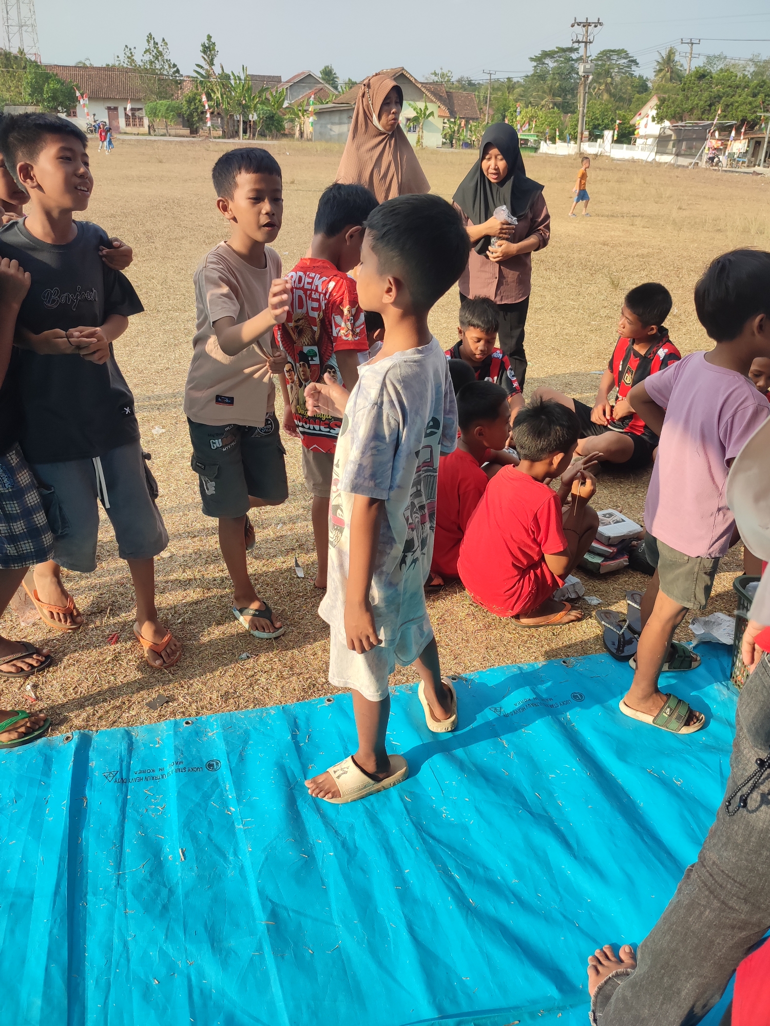 The image depicts a group of Indonesian children and two adult women gathered outdoors on a dry, grassy field in Nusa Jaya, Indonesia. The children, ranging in age from roughly six to twelve, are dressed in casual clothing, including t-shirts and shorts. Some of the children are standing, while others are sitting or crouching on a bright blue tarp spread on the ground. The adults, both wearing headscarves, appear to be supervising the children. One woman in a brown shirt and headscarf is holding a plastic bottle, while the other, in a black hijab, is also holding something in her hand.
In the background, houses with red and white flags are visible, suggesting a local community or event. The landscape is dry and open, with a few trees and utility poles in the distance. The overall atmosphere appears to be a casual gathering or activity, possibly related to a school event or community outreach program, given the presence of the children and adults in an open field. The lighting suggests it is daytime, likely late afternoon given the angle of the sun. The image is taken from a slightly elevated perspective, looking down on the group.