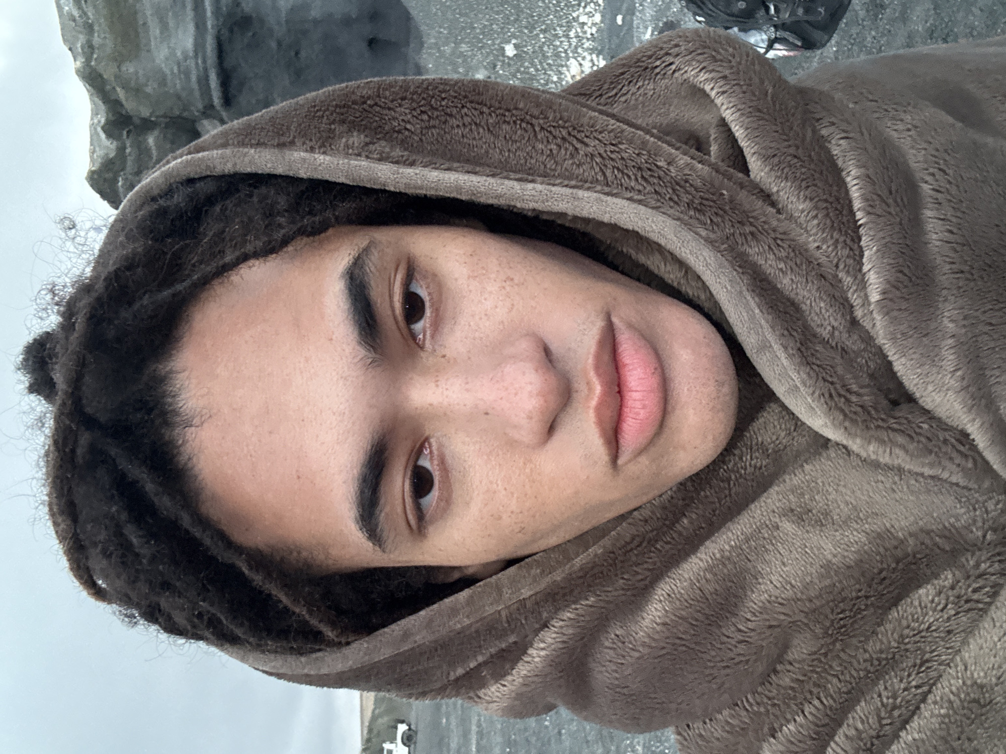 A person with dark brown dreadlocks, wearing a fuzzy brown hooded jacket, is seen in a close-up shot. The individual has brown eyes, freckles, and full lips. The background includes what appears to be a rocky area and a body of water under an overcast sky. The location could be near a coastal area in Teguise, Spain, but without additional context, this cannot be conclusively determined. A small white vehicle is visible in the distance at the bottom of the image.